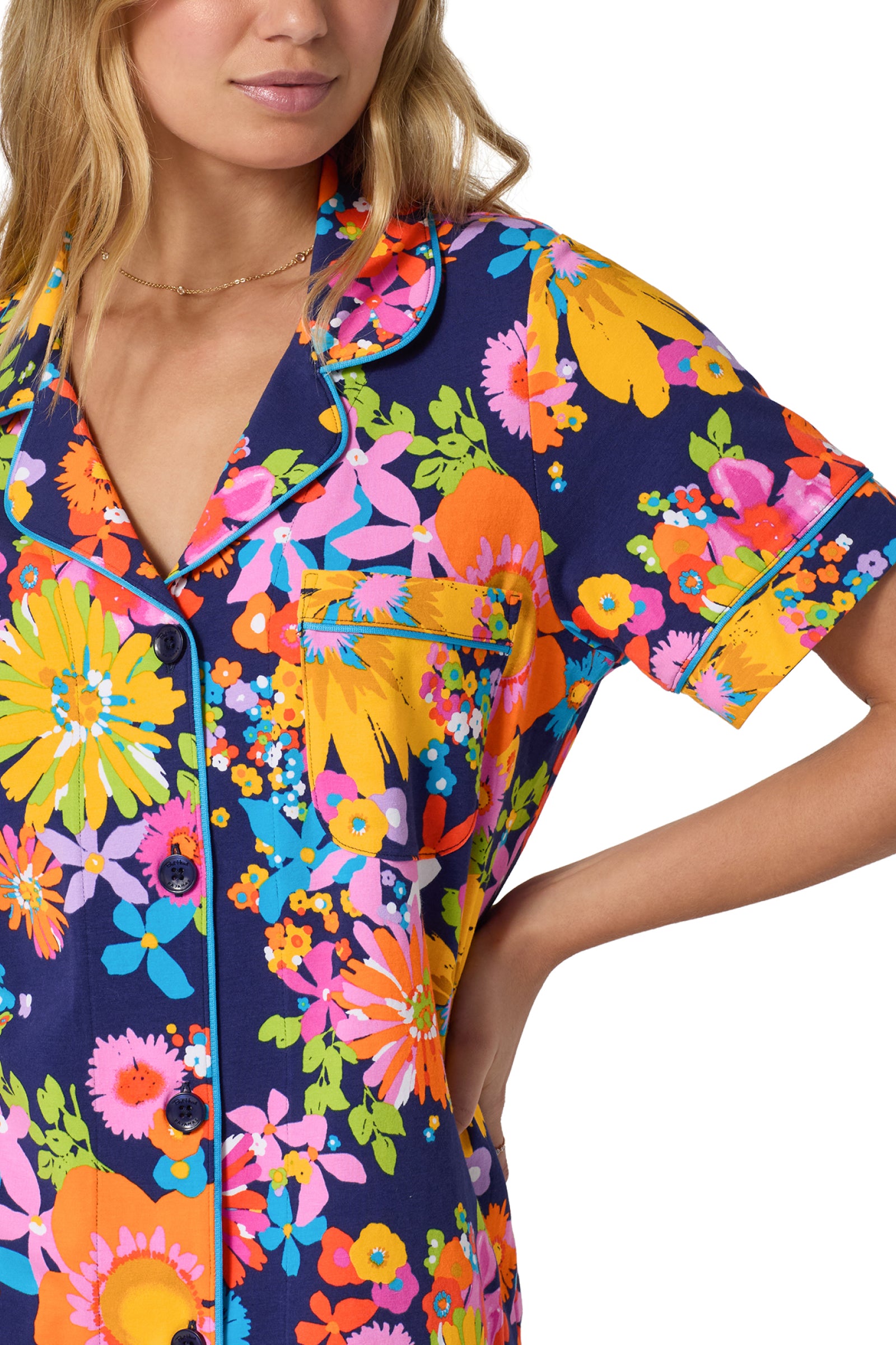 A woman wears the Shorty PJ Set - Bay St Bloom: a navy blue short-sleeve pajama top with yellow, orange, pink, and green floral print, collar, buttons, and chest pocket. Her hand is on her hip.