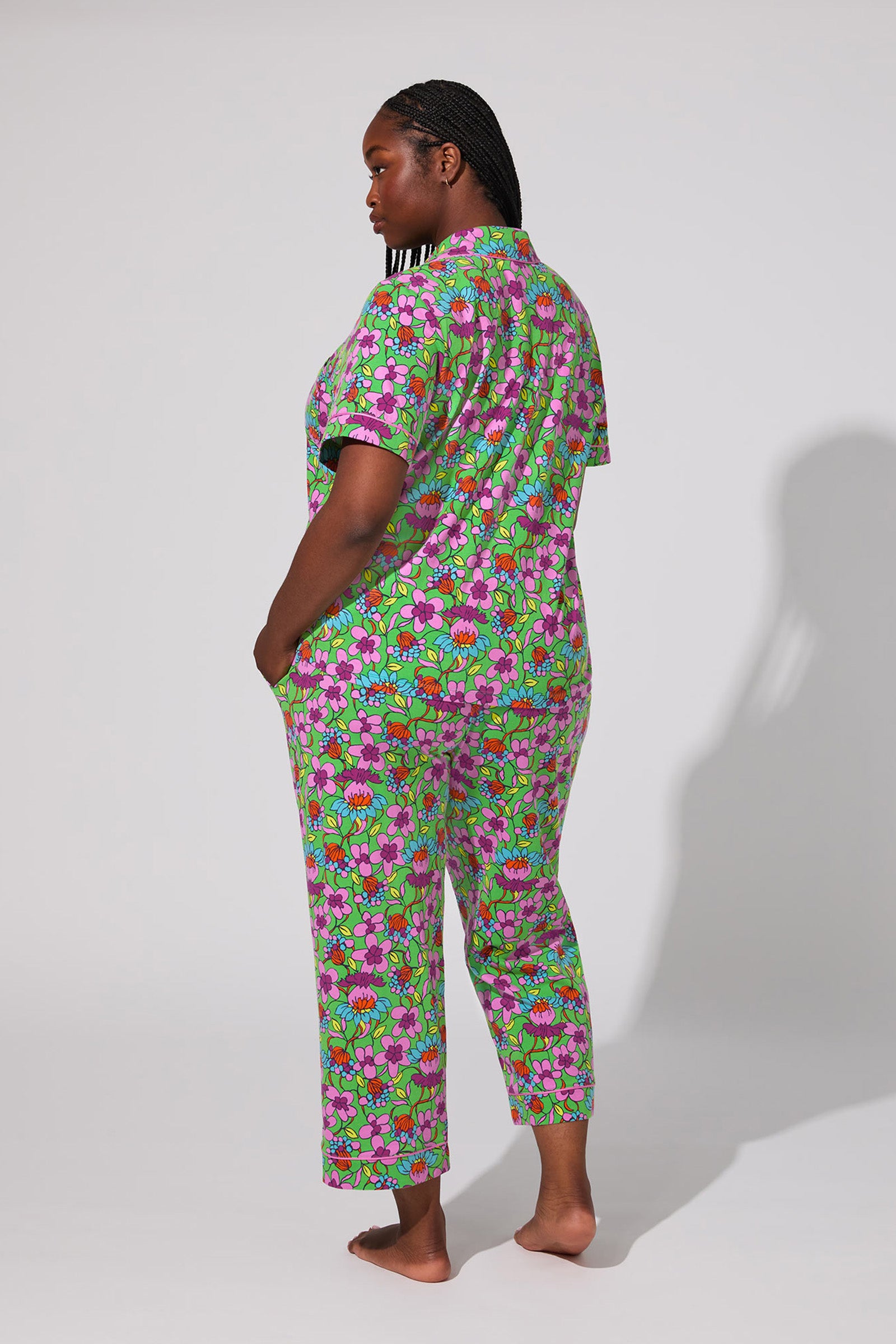 A woman stands barefoot, facing slightly away, wearing the Crop Classic PJ Set - Garden Floral, a colorful short-sleeve pajama set with bright green, pink, and purple floral patterns against a plain background.