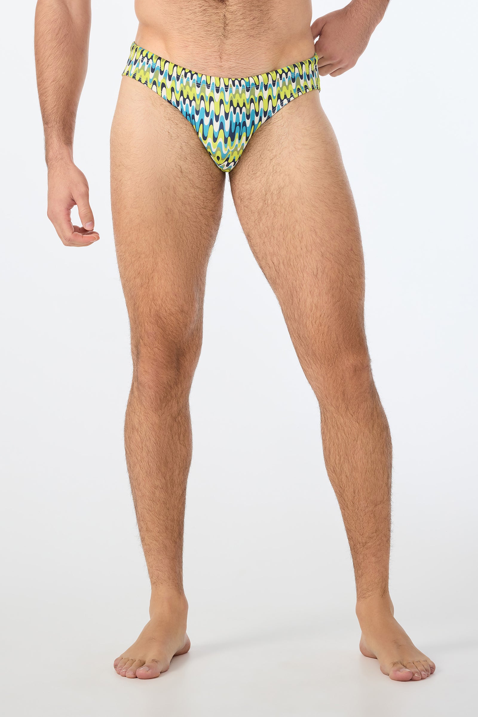 A man is barefoot, modeling the Lagos Swim Brief - Psychedelic Voyage, featuring a green, blue, white, and yellow zigzag design. His arms and face are partially out of frame against a plain white background.