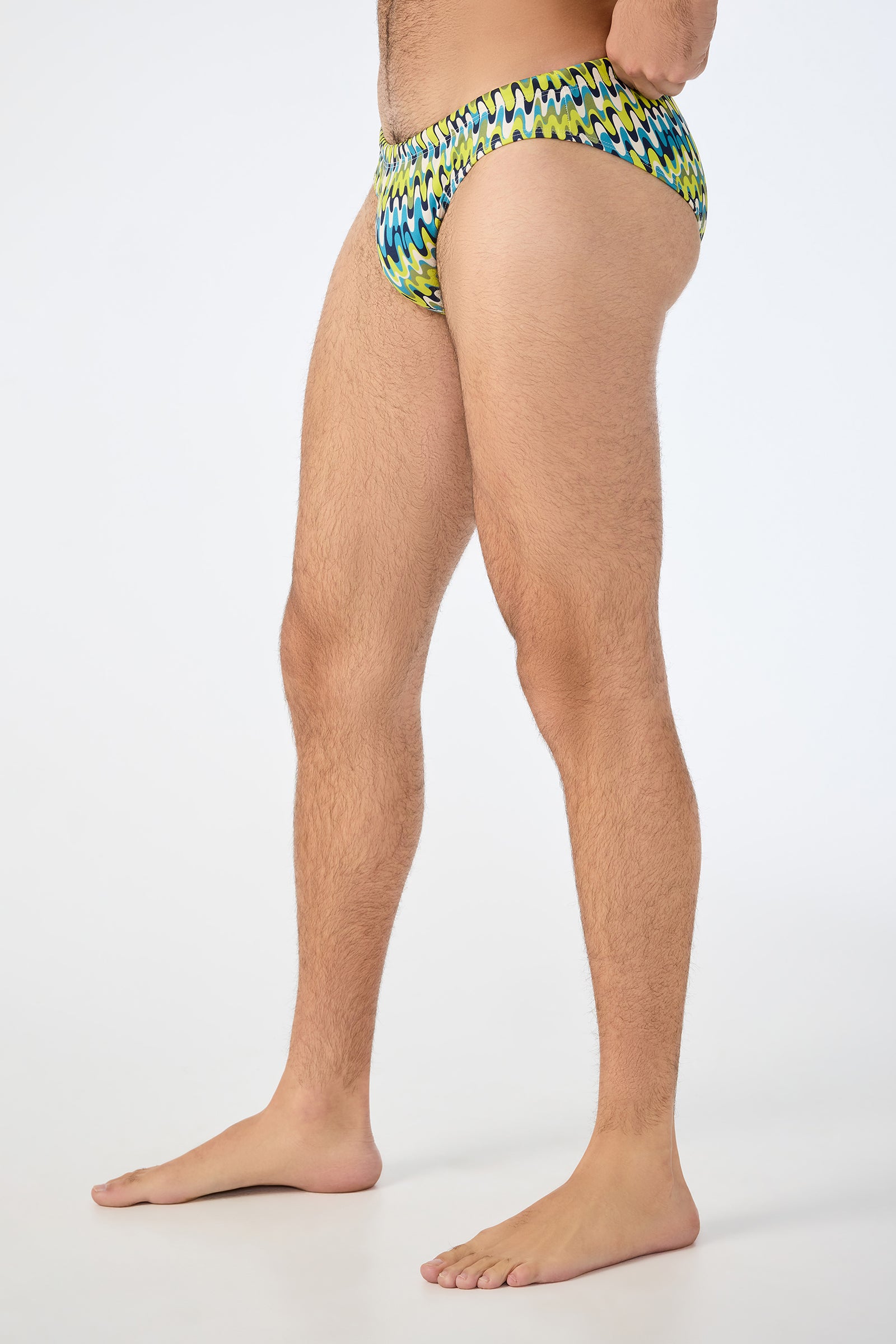 A man stands barefoot in profile against a plain white background, wearing the Lagos Swim Brief - Psychedelic Voyage with a vibrant zigzag pattern. His face is not visible.