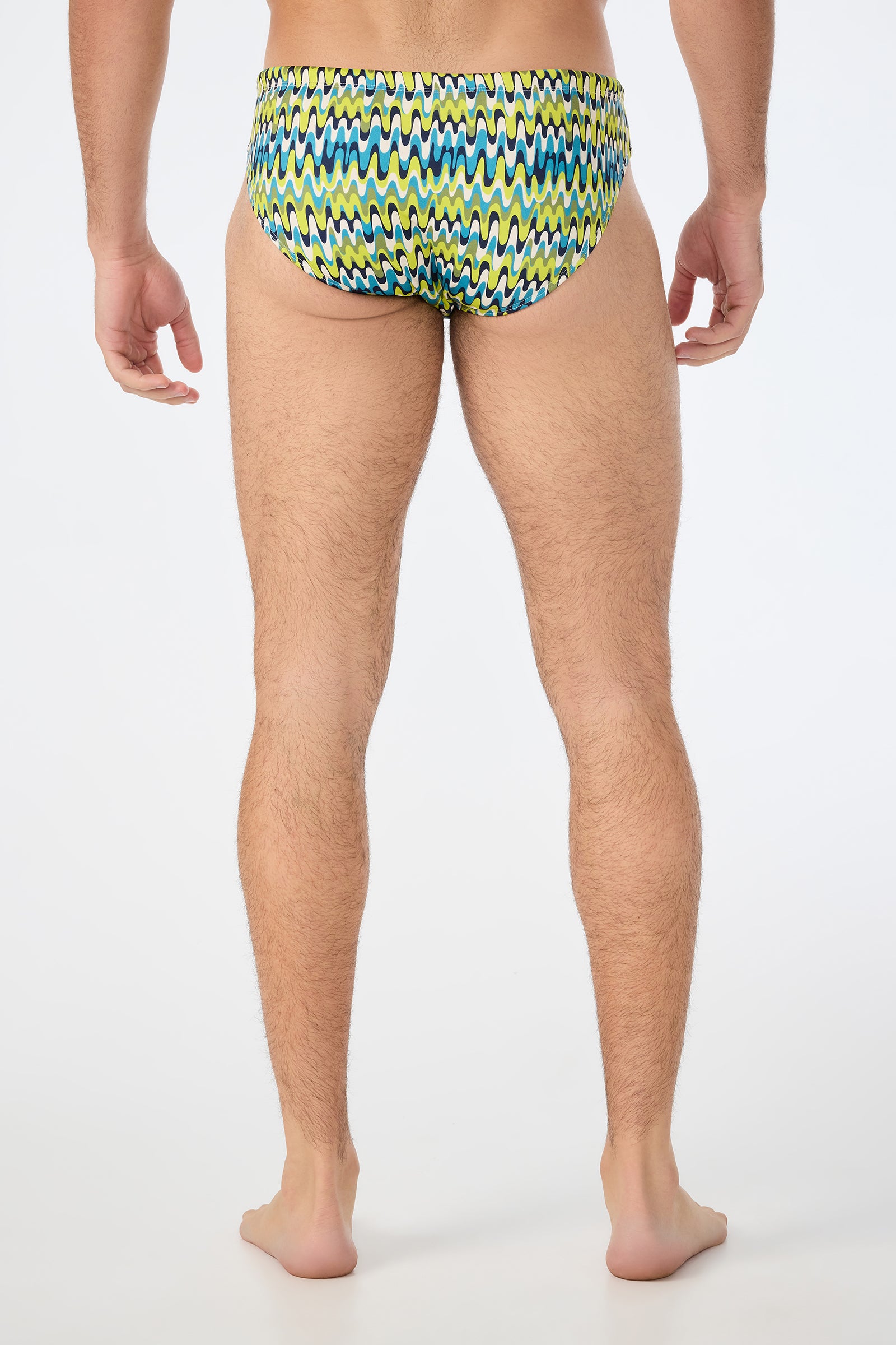 A man stands barefoot with his back to the camera, wearing the Lagos Swim Brief - Psychedelic Voyage featuring a blue, yellow, and green zigzag pattern on a white background.