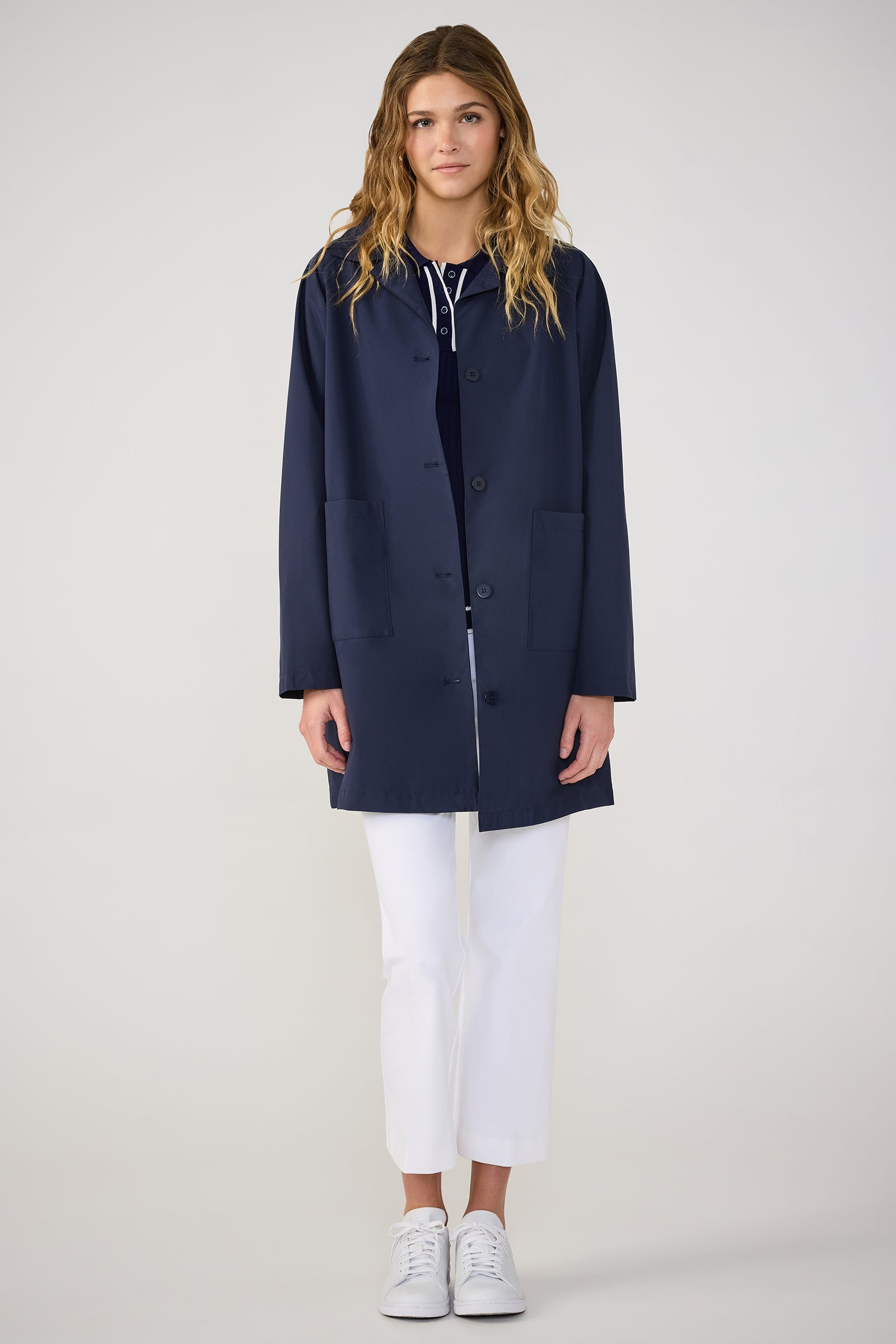 A person with long wavy hair stands against a plain light background, facing forward with arms relaxed, wearing the TT X SR Mac Rain Coat in Ink, along with white pants and white sneakers.