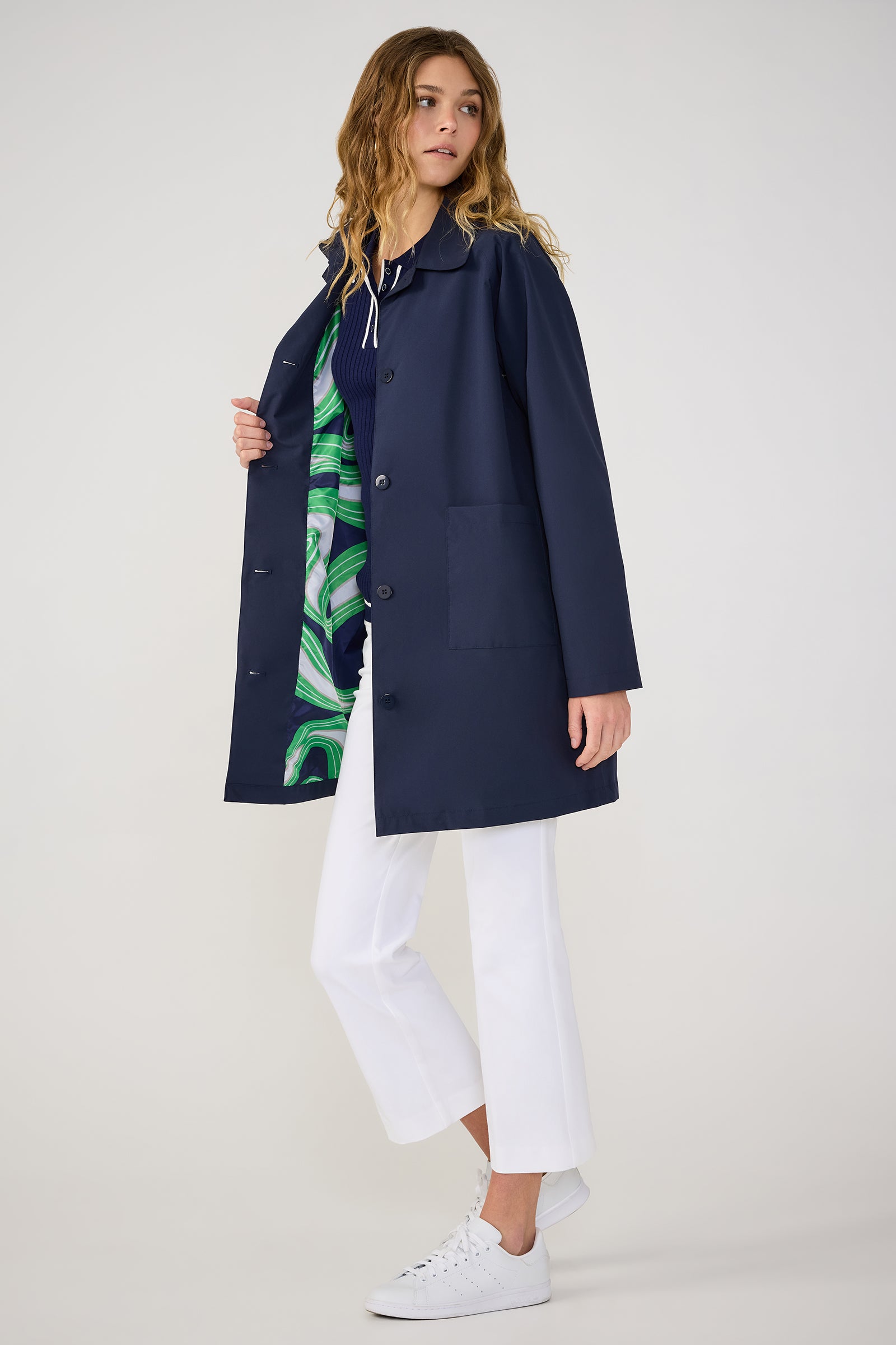 A woman poses against a plain, light background in the TT X SR Mac Rain Coat in Ink, worn open over a patterned green and blue top, paired with white cropped pants and white sneakers. She has wavy hair.