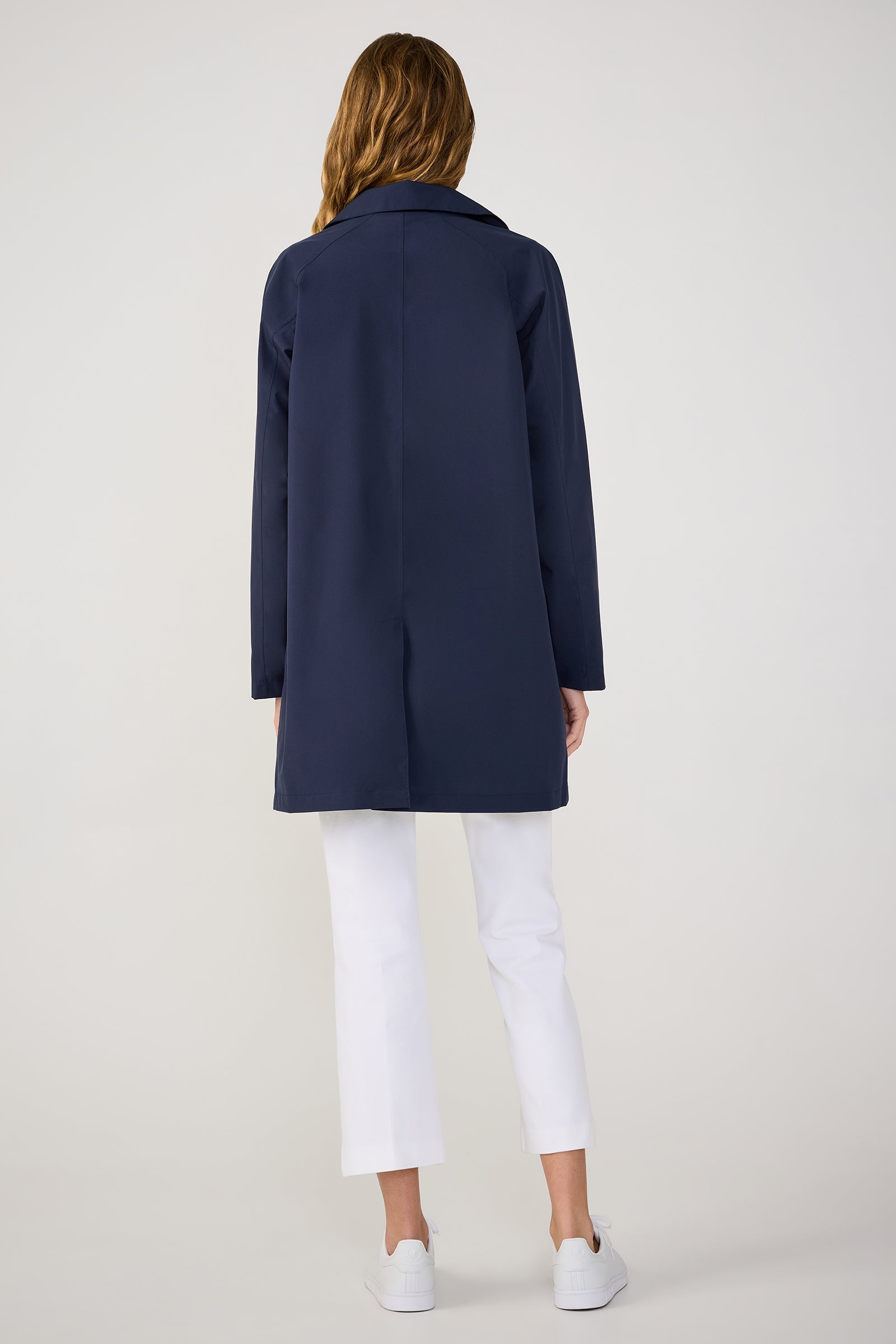A woman faces away, wearing the TT X SR Mac Rain Coat in Ink, paired with white cropped pants and white sneakers against a plain light background.