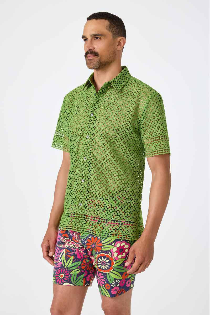 A man with short hair and a mustache wears the Belmont Shore Shirt, a sheer green short-sleeve button-up with a geometric cutout pattern, paired with colorful floral shorts. He stands against a plain white background.