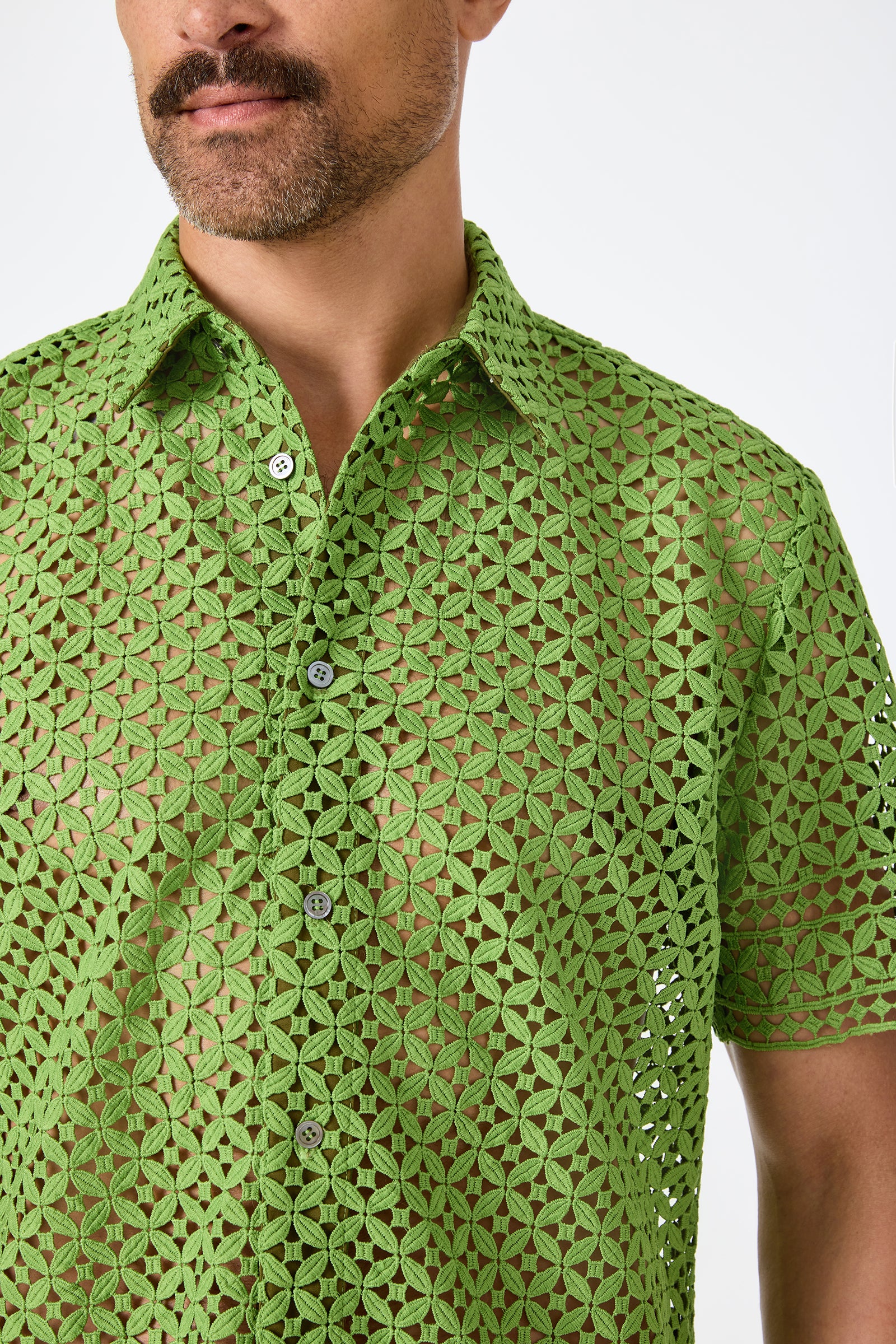A man wears the Belmont Shore Shirt—a short-sleeved, button-up green top crafted from see-through geometric lace. Only the lower part of his face and torso are visible against a plain background.