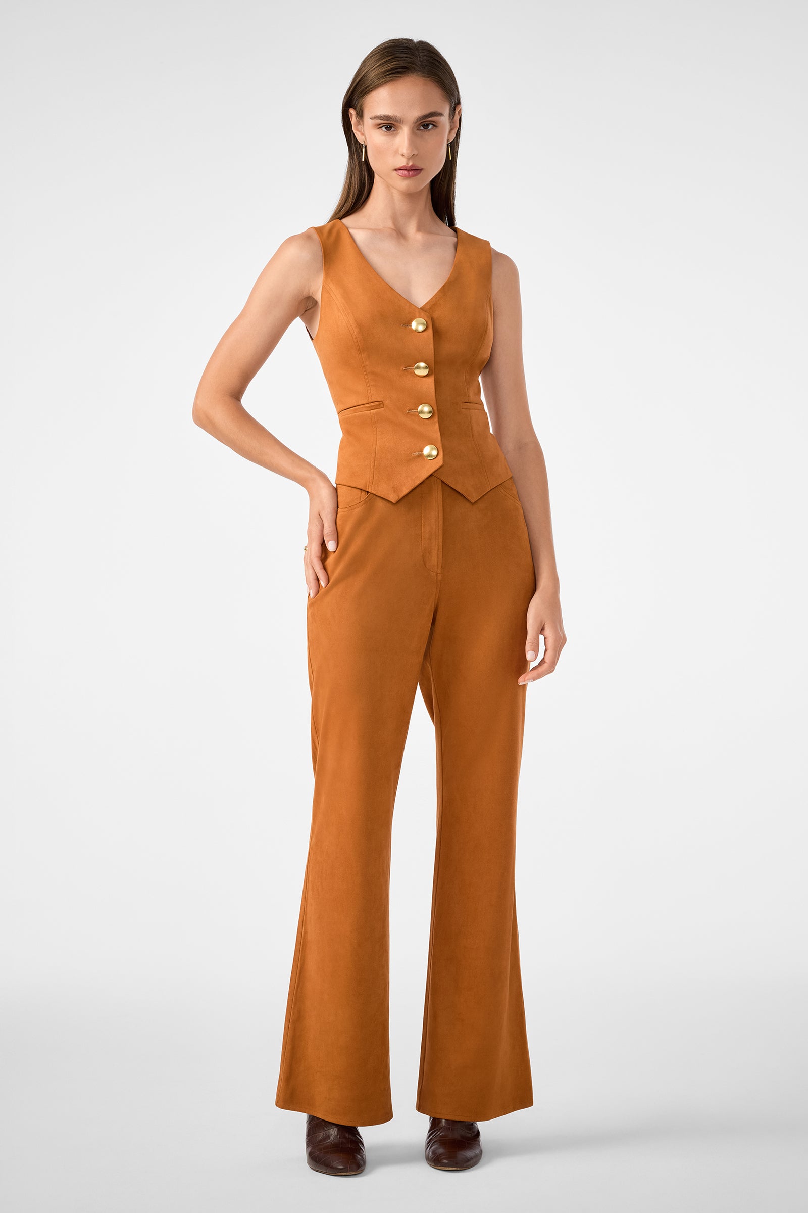 A woman faces forward wearing the Kofi 2 Vest—a sleeveless, fitted tan vest with large gold buttons—and matching high-waisted, flared pants. She has straight brown hair, brown shoes, and stands against a plain light background.