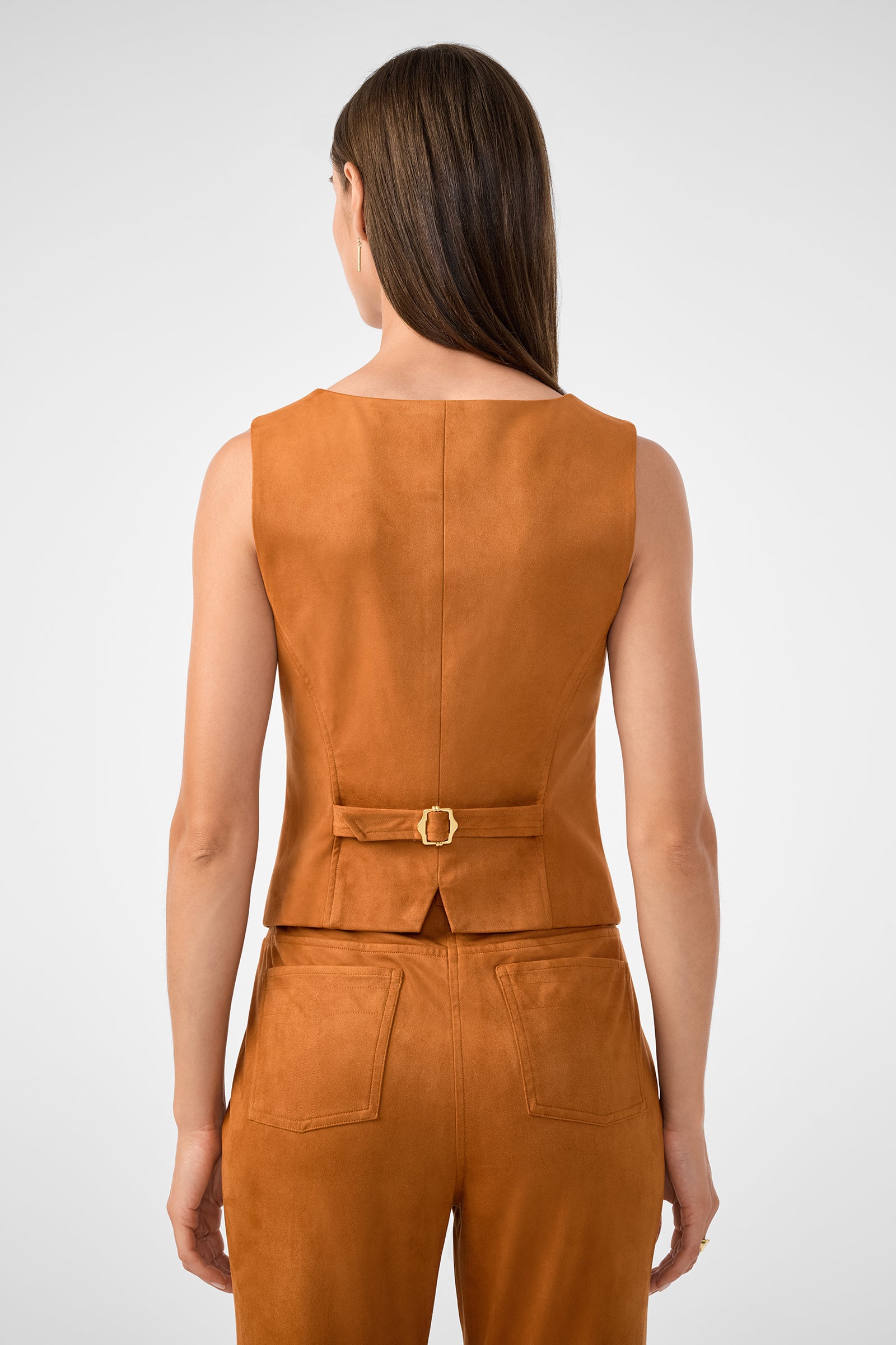 A woman with straight brown hair is seen from behind wearing the Kofi 2 Vest, a sleeveless rust suede vest with a small back belt featuring a gold buckle and a short slit at the hem, paired with matching pants.