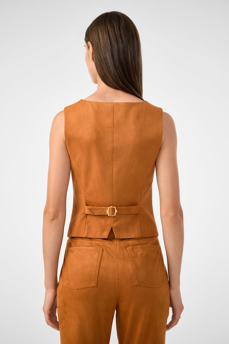 A woman with straight brown hair is seen from behind wearing the Kofi 2 Vest, a sleeveless rust suede vest with a small back belt featuring a gold buckle and a short slit at the hem, paired with matching pants.