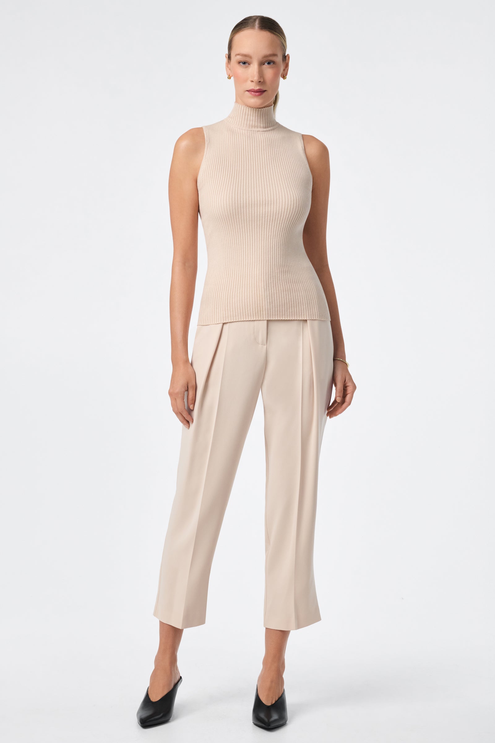 A woman with slicked-back hair and a neutral expression stands against a white background wearing the Westhampton Knit Pullover, a sleeveless beige ribbed mock-neck top, paired with beige ankle-length pants and black heeled shoes.