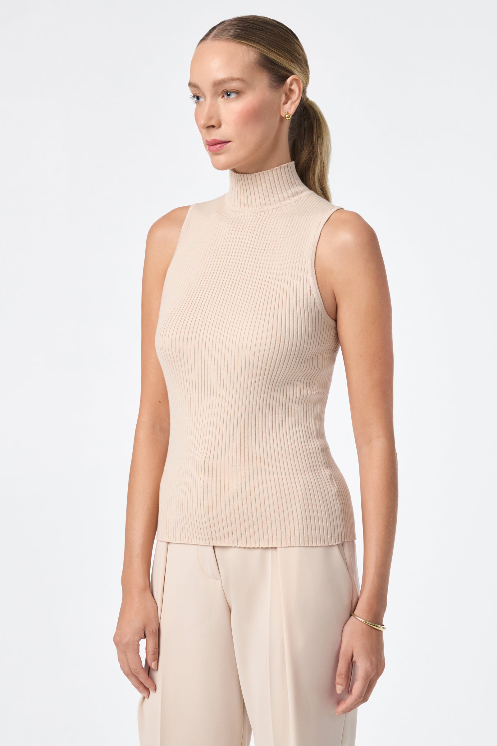 A woman with light skin and straight, pulled-back hair models the sleeveless beige Westhampton Knit Pullover with matching pants against a plain white background.