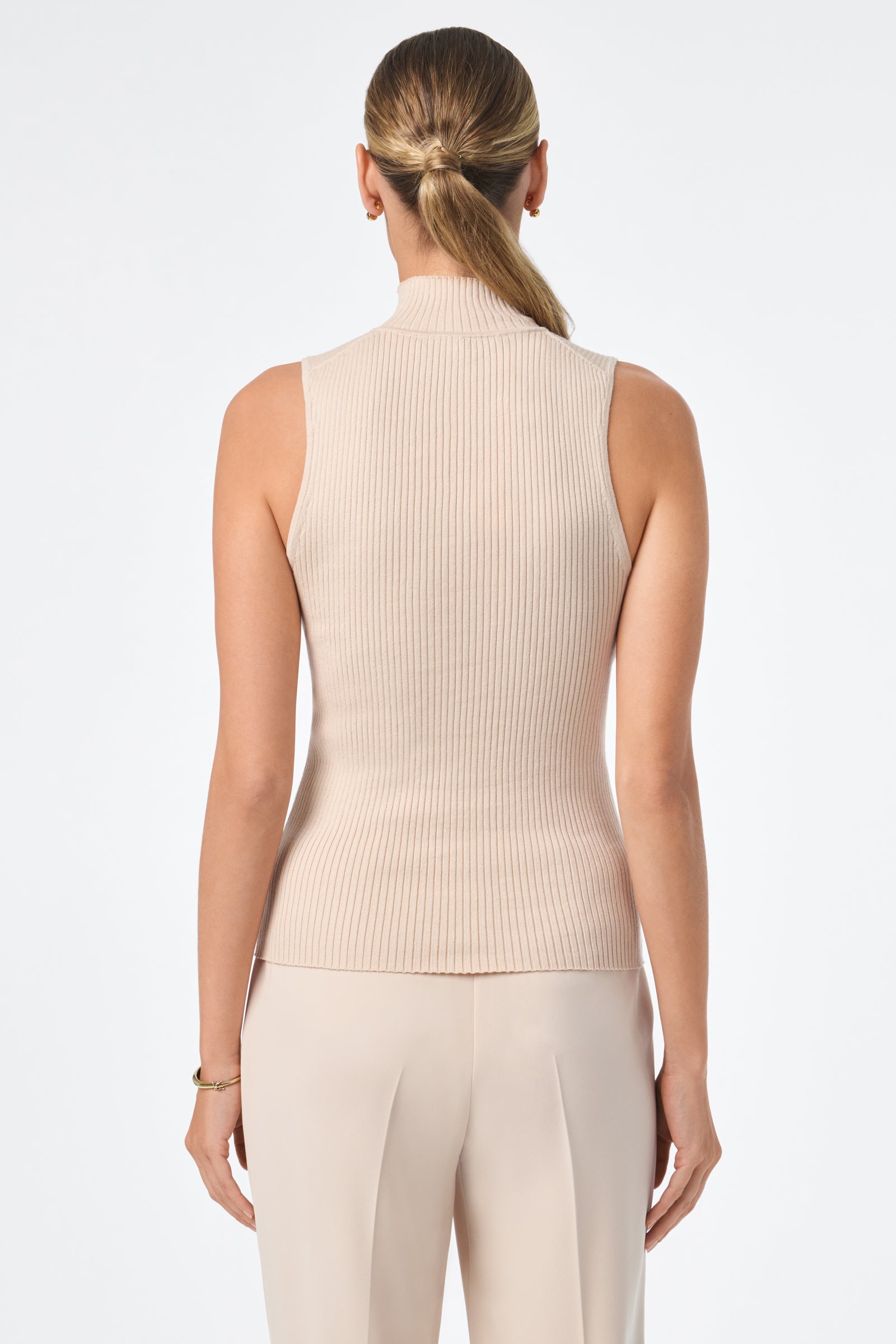 A woman with her hair in a low ponytail faces away, wearing the Westhampton Knit Pullover—a sleeveless, beige ribbed turtleneck top—paired with light pants. The background is plain white.