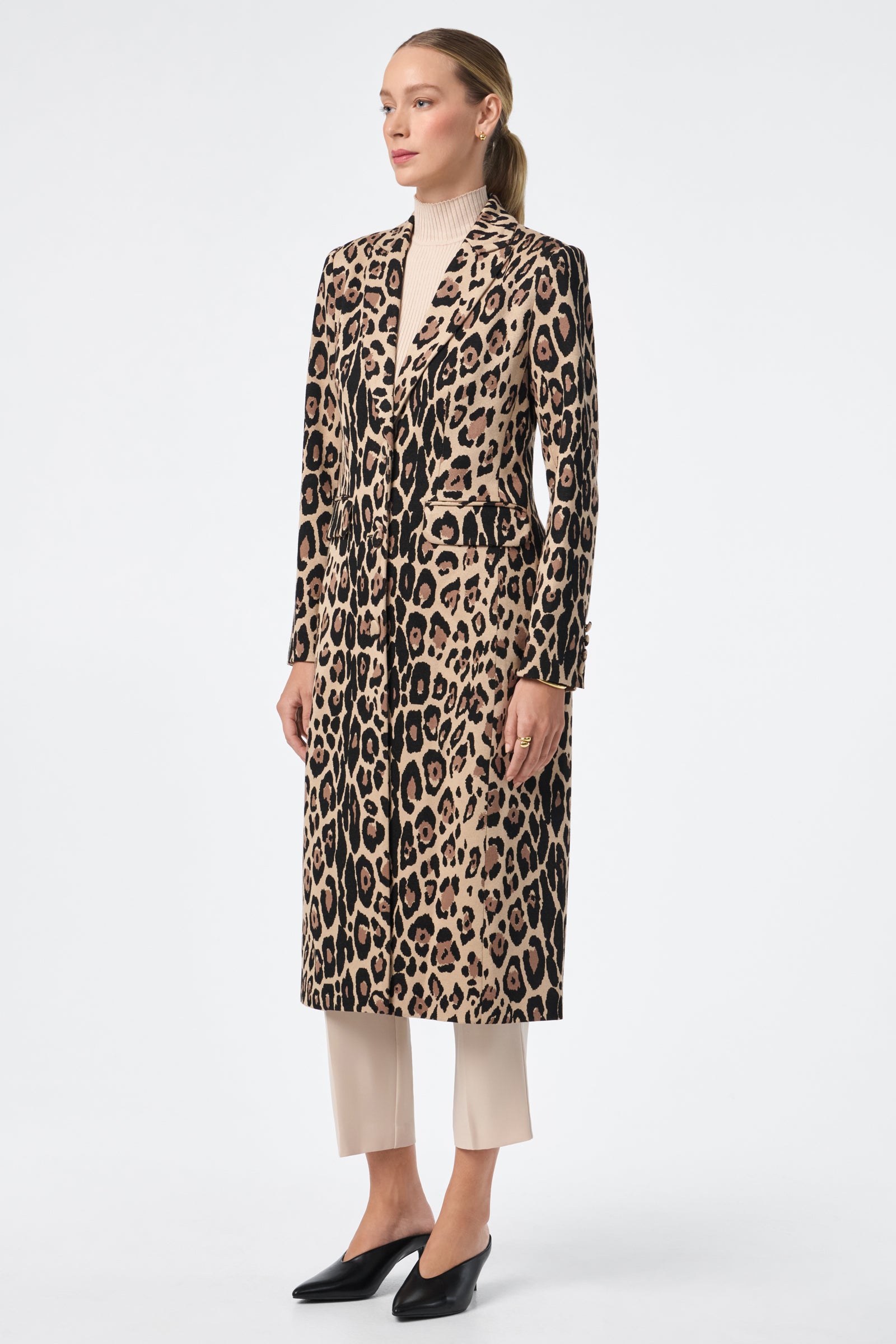 A woman faces left, wearing the Playbill Coat—a long, leopard-print piece—over a beige turtleneck and cream pants with black shoes. Her hair is tied back in a low ponytail against a plain white background.