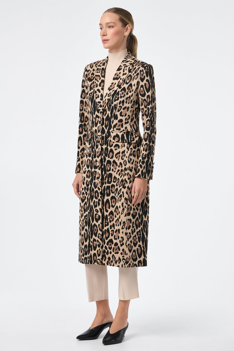 A woman faces left, wearing the Playbill Coat—a long, leopard-print piece—over a beige turtleneck and cream pants with black shoes. Her hair is tied back in a low ponytail against a plain white background.