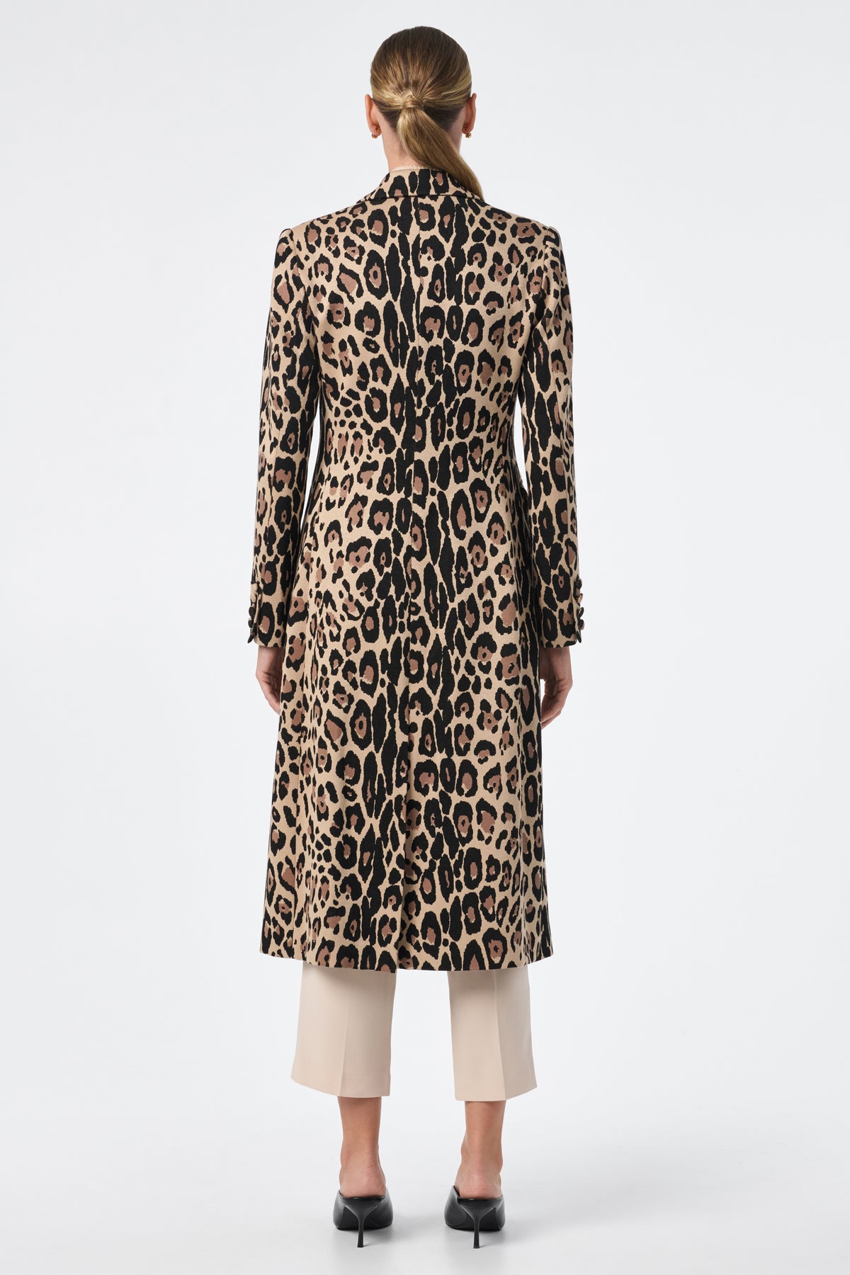 A person with long hair tied back is shown from behind, wearing the Playbill Coat—a long, fitted leopard print piece—over beige cropped pants and black low-heeled shoes, against a plain white background.