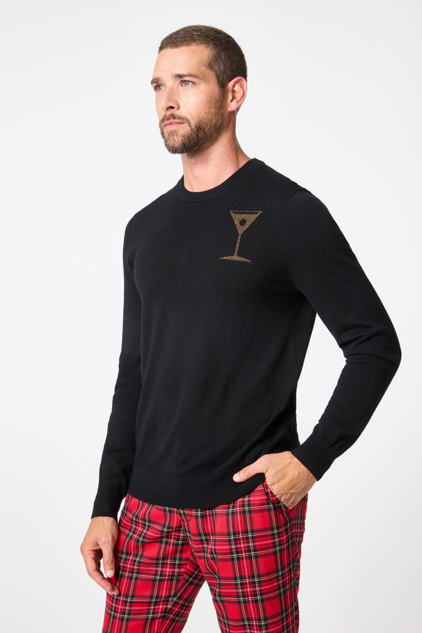 A man with short hair and a beard models the Martini Crewneck Sweater, featuring a martini glass design on the chest, paired with red plaid pants. He stands against a plain white background, one hand in his pocket, looking to the side.