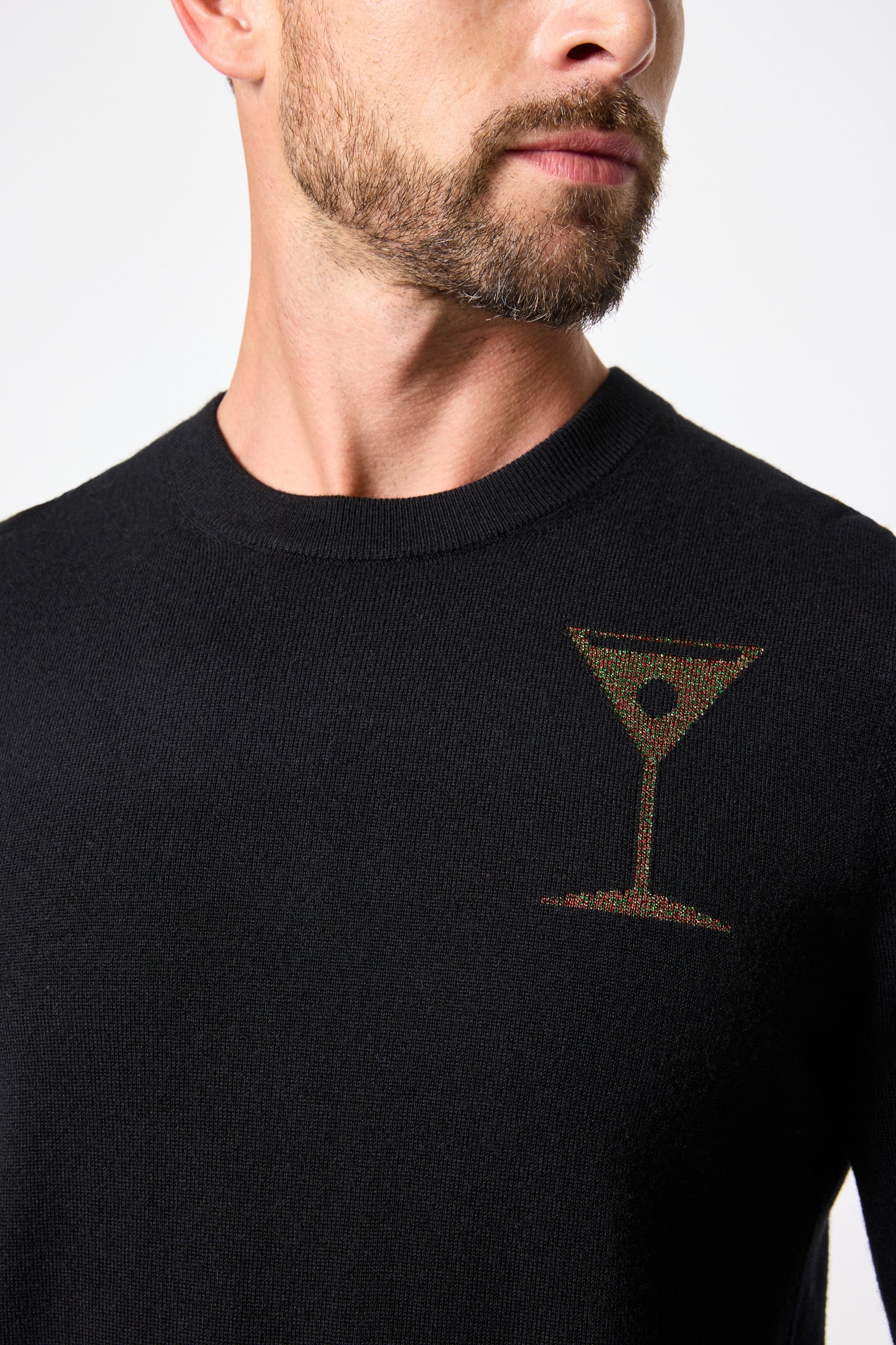 A man models the Martini Crewneck Sweater—black with a small embroidered martini glass on the chest—shown from neck to mid-torso, facing slightly sideways.