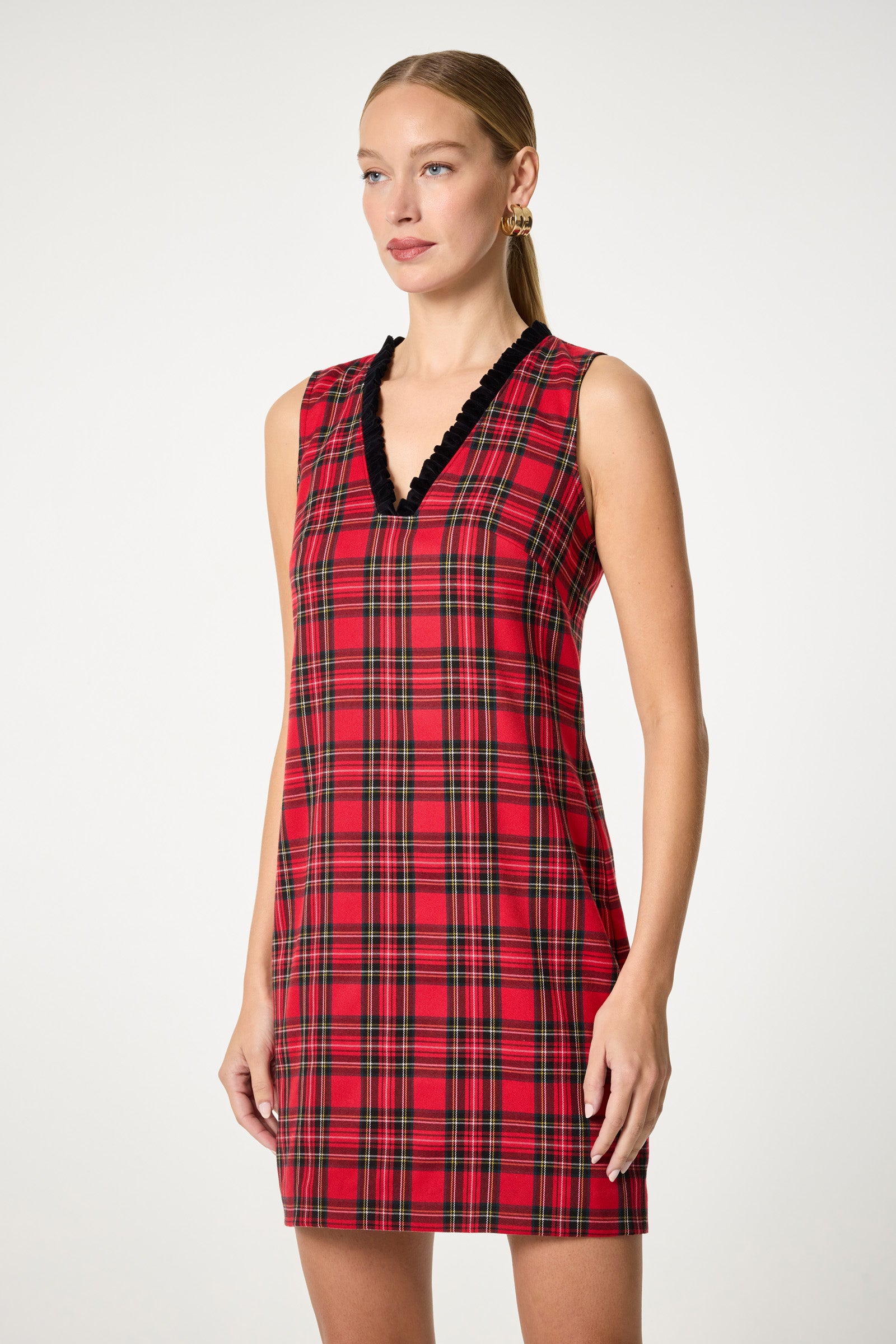 A woman with light skin and straight blonde hair pulled back models the Revah Tartan Mini Dress—a sleeveless, red plaid design with a black v-neck trim—against a plain light background. She accessorizes with gold earrings.