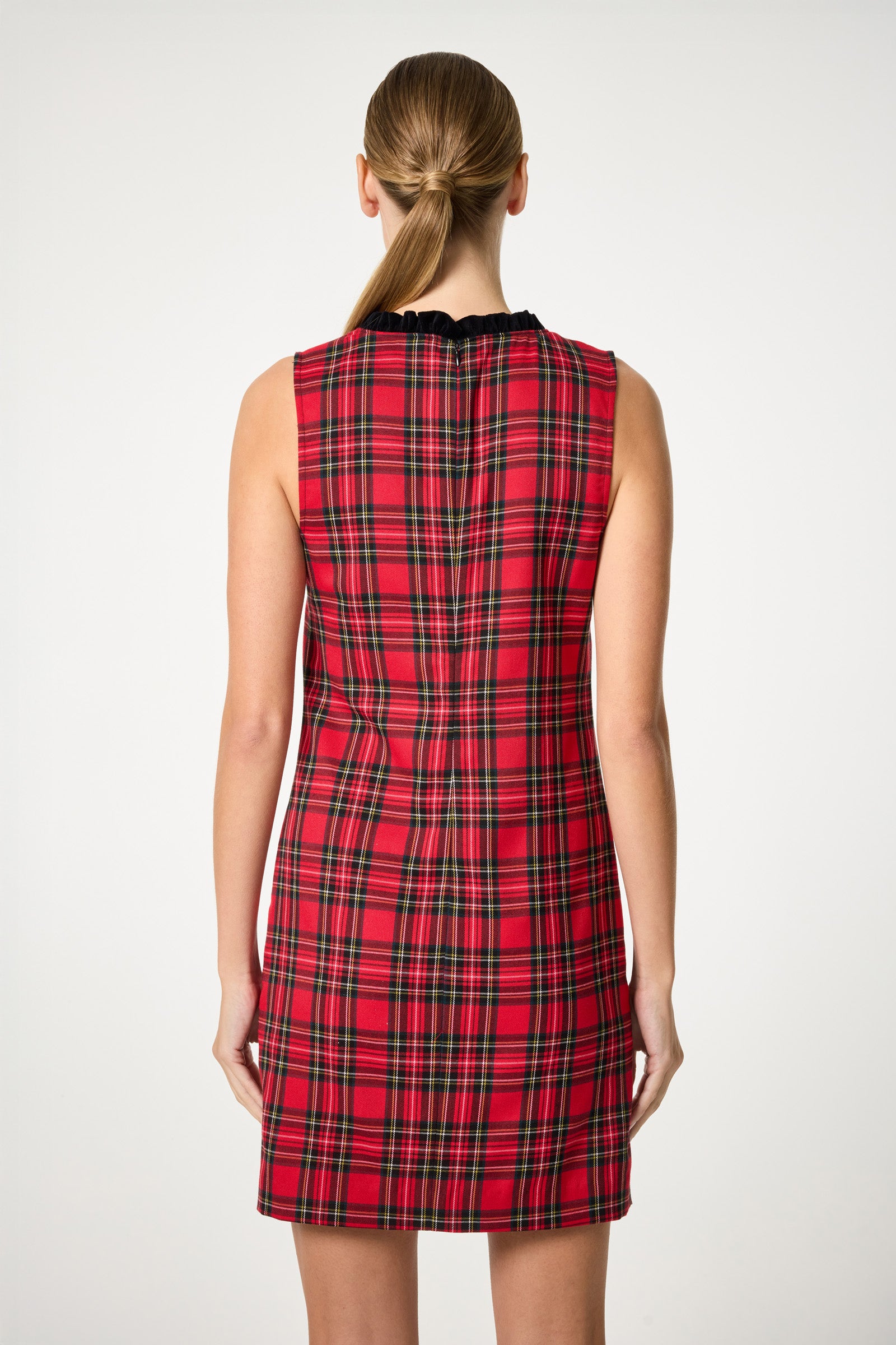 A woman with long, straight hair stands with her back to the camera against a plain, light background, wearing the sleeveless, knee-length Revah Tartan Mini Dress in red plaid.