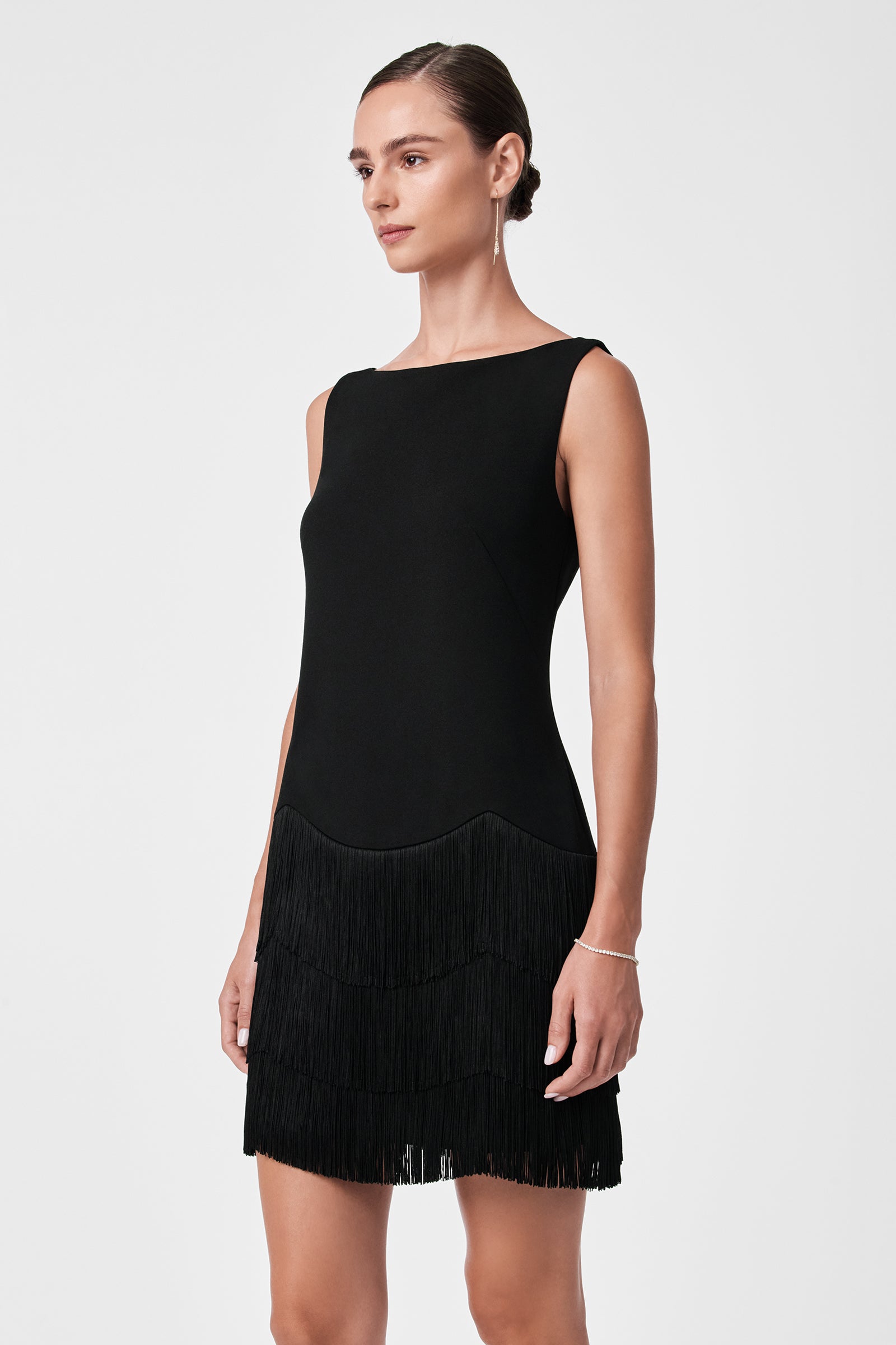 A woman in the Palais Dress—a sleeveless black dress with tiered fringe on the skirt—stands against a plain white background, gazing slightly to the side.
