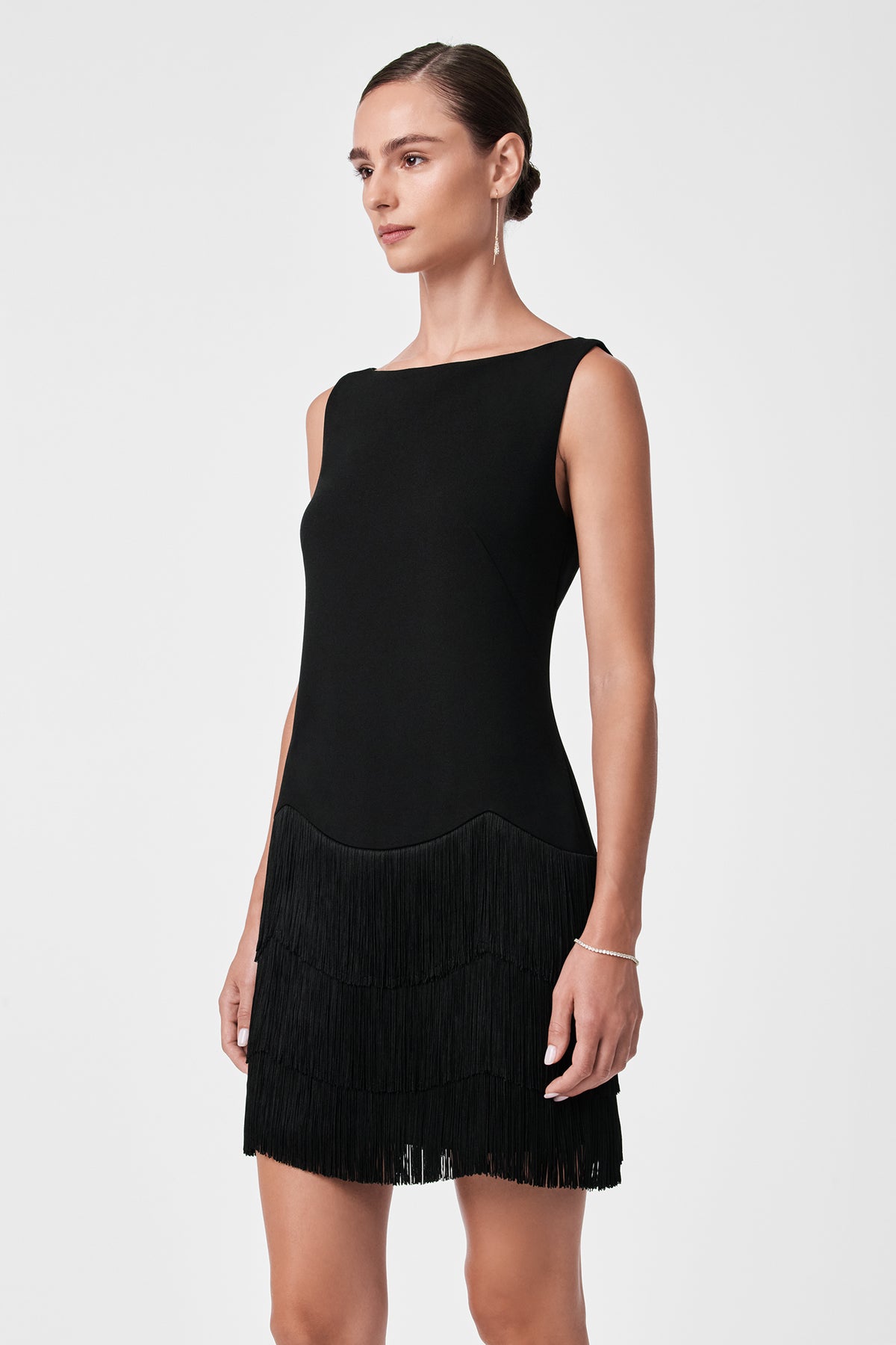 A woman in the Palais Dress—a sleeveless black dress with tiered fringe on the skirt—stands against a plain white background, gazing slightly to the side.