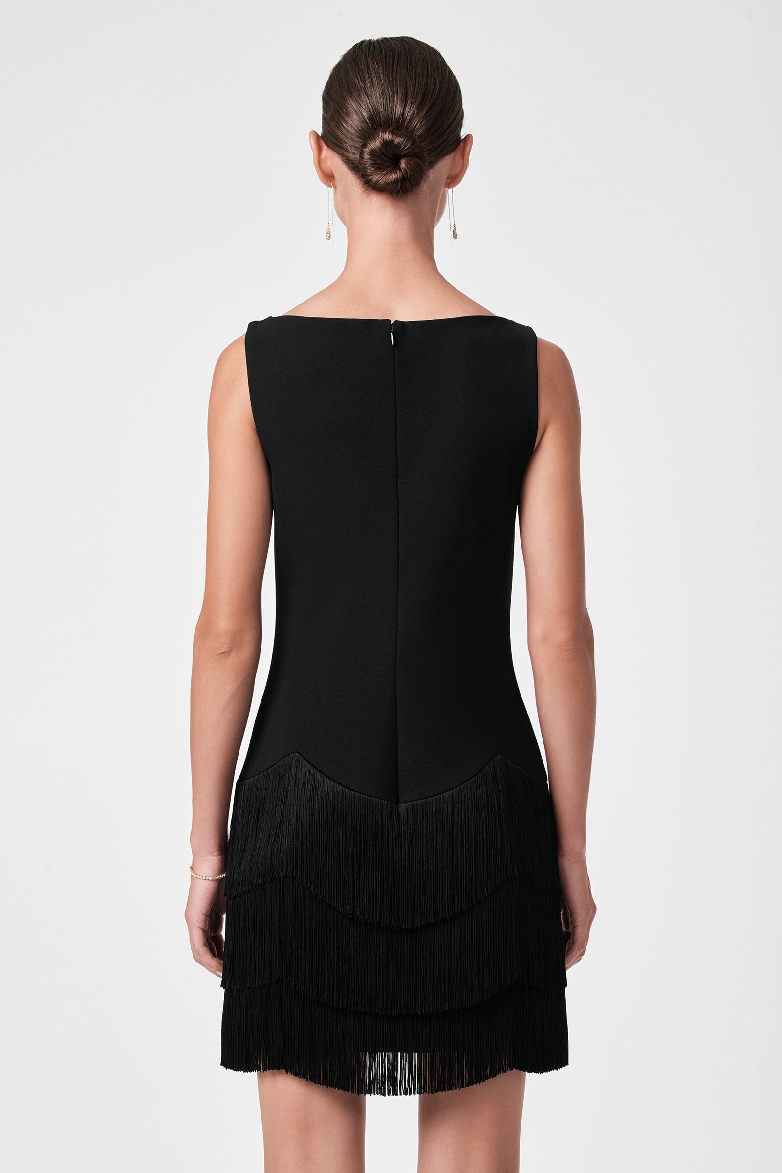 A woman with her hair in a neat bun is seen from behind, wearing the Palais Dress—a sleeveless black dress with a layered fringe skirt—and simple drop earrings, standing against a plain light background.