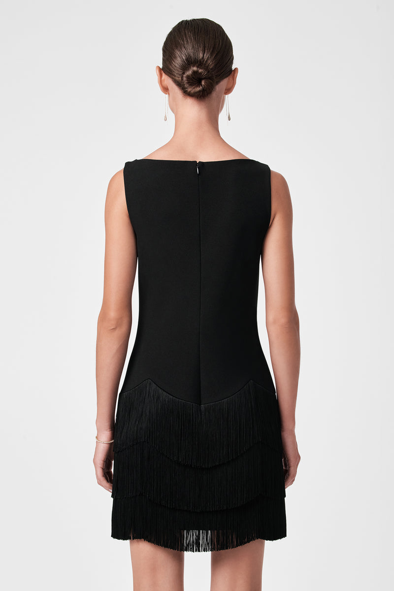 A woman with her hair in a neat bun is seen from behind, wearing the Palais Dress—a sleeveless black dress with a layered fringe skirt—and simple drop earrings, standing against a plain light background.