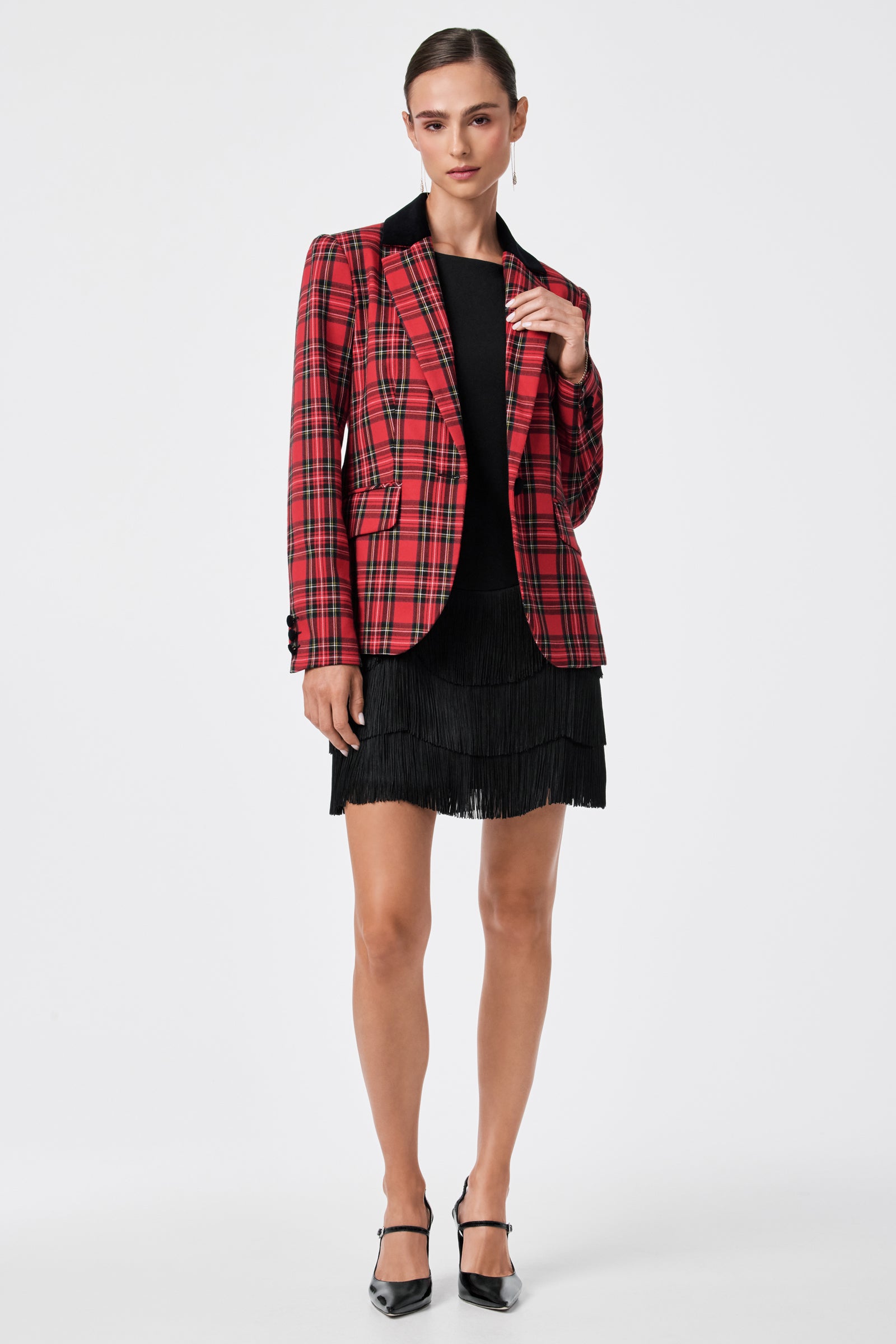 A person stands against a plain white background wearing the Palais Dress under a red plaid blazer and black heels, with hair pulled back and one hand touching the blazer’s lapel.