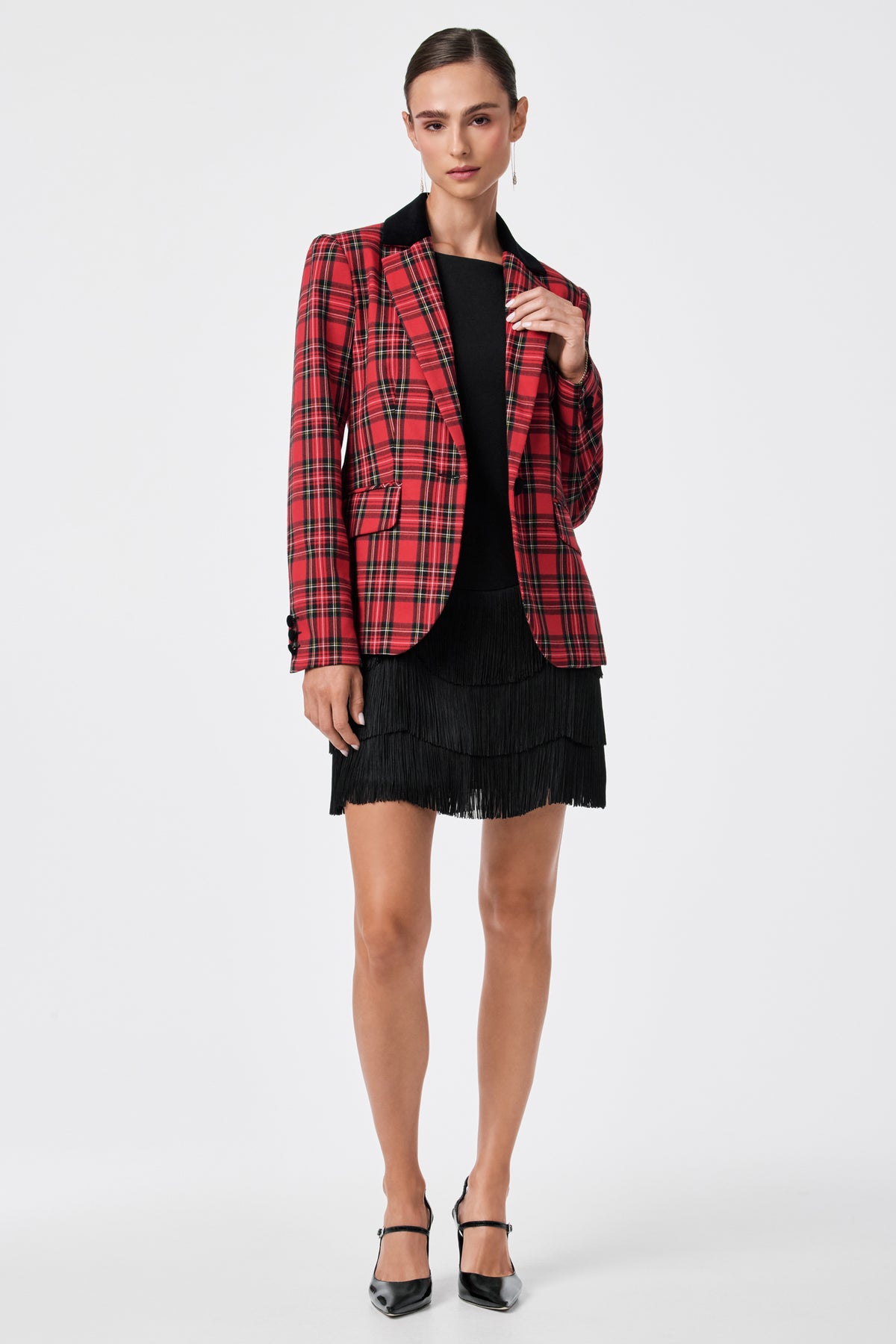 A person stands against a plain white background wearing the Palais Dress under a red plaid blazer and black heels, with hair pulled back and one hand touching the blazer’s lapel.