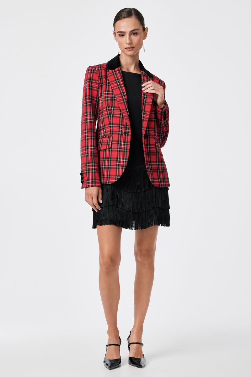 A person stands against a plain white background wearing the Palais Dress under a red plaid blazer and black heels, with hair pulled back and one hand touching the blazer’s lapel.