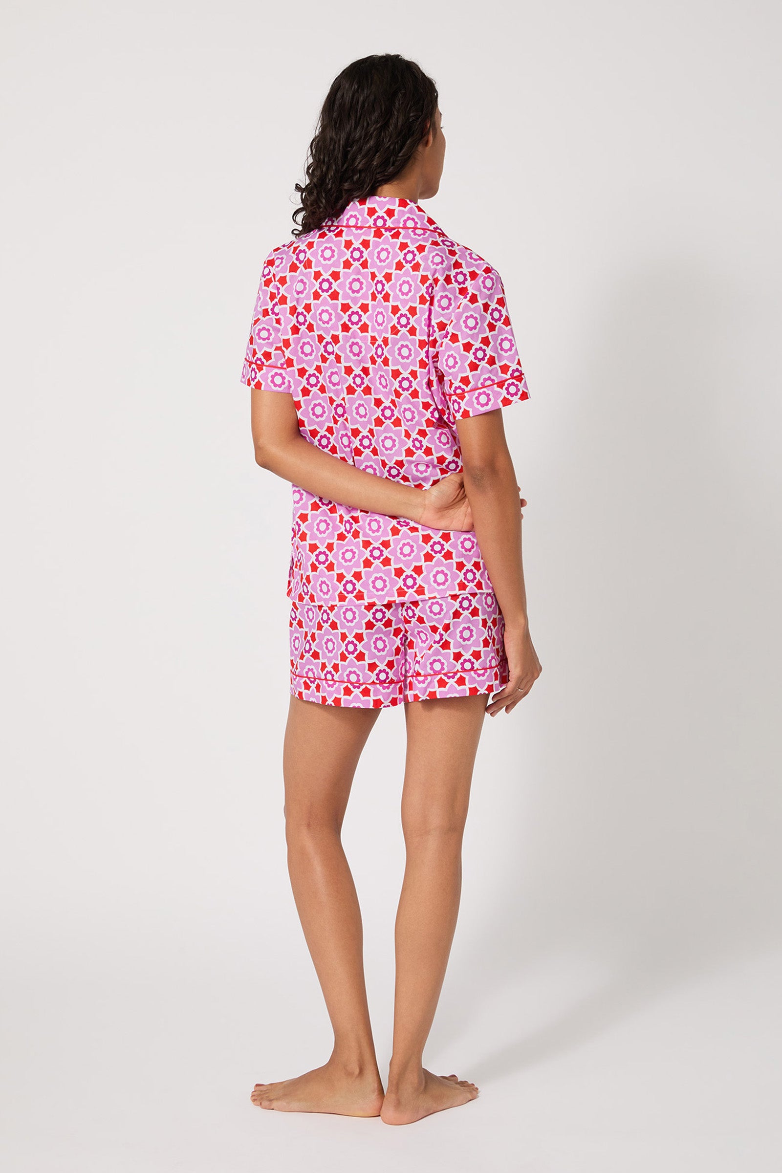 A person stands barefoot, facing away, wearing the Shorty PJ Set - Mosaic Tile featuring a pink, red, and white geometric pattern. One arm is bent behind their back against a plain white background.