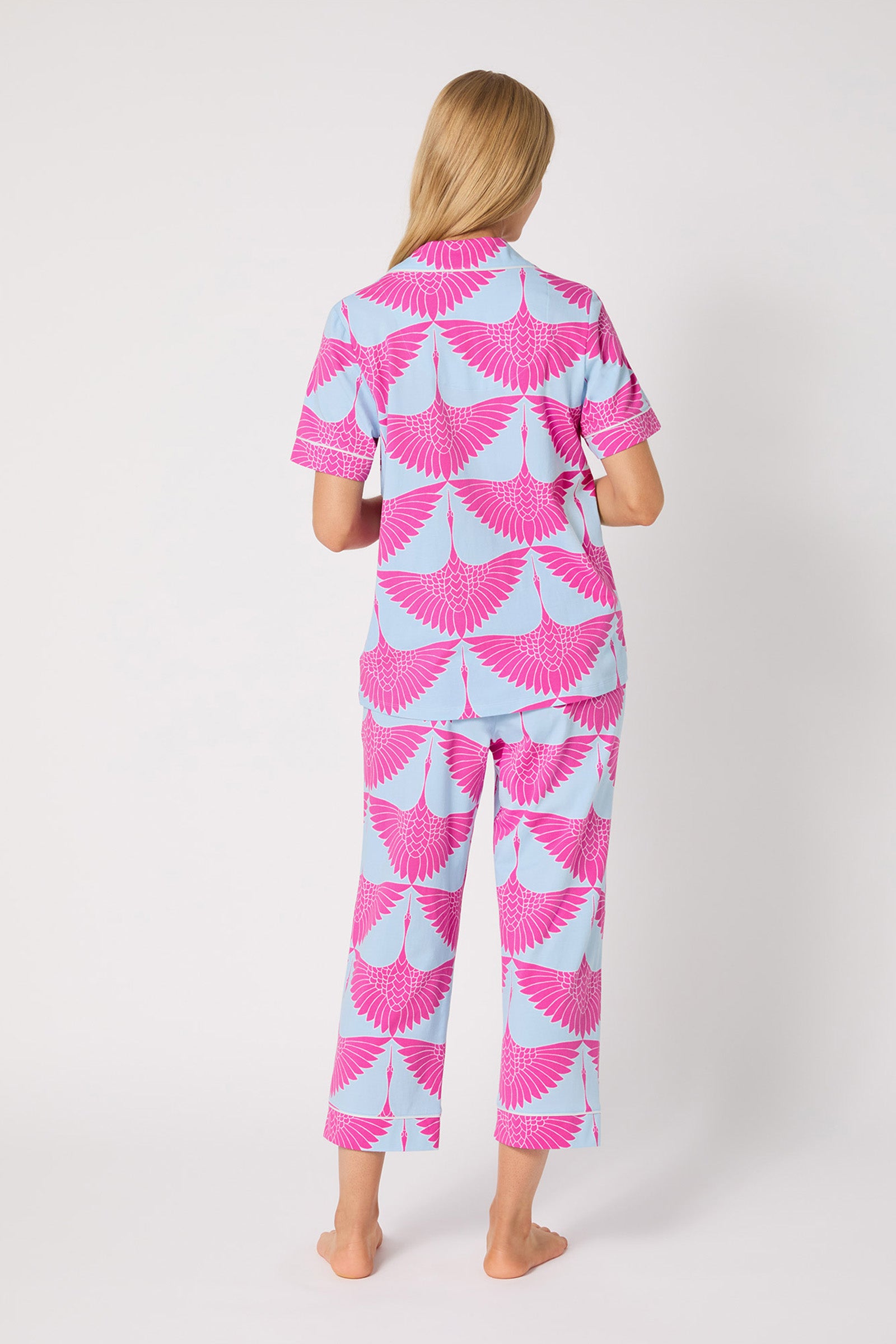 A person with long blonde hair stands barefoot facing away, wearing the Crop PJ Set - Cranes, featuring matching light blue pajamas with a bold pink crane wing pattern on both the top and pants. The background is plain white.