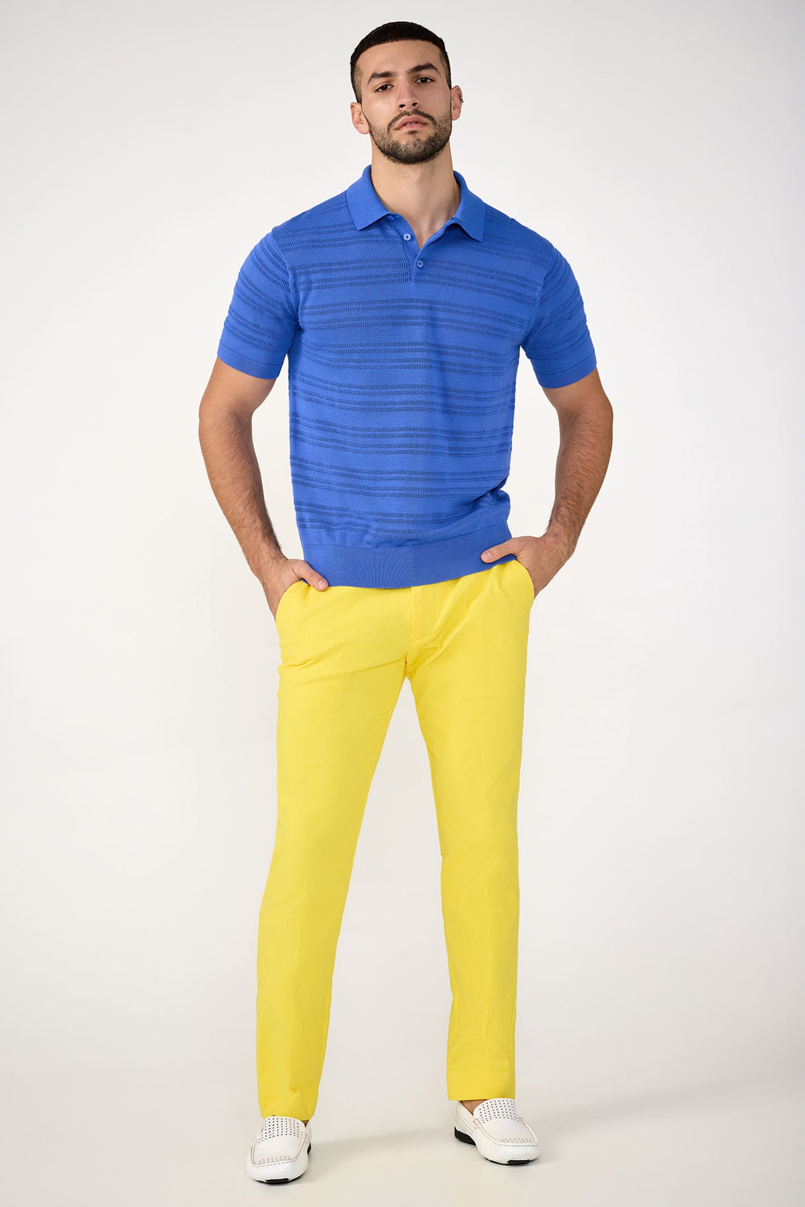 A man stands against a plain background wearing a bright blue polo, white shoes, and the Clyde Slim Trouser - Stretch Herringbone in yellow. His hands are in his pockets as he faces forward with a neutral expression.
