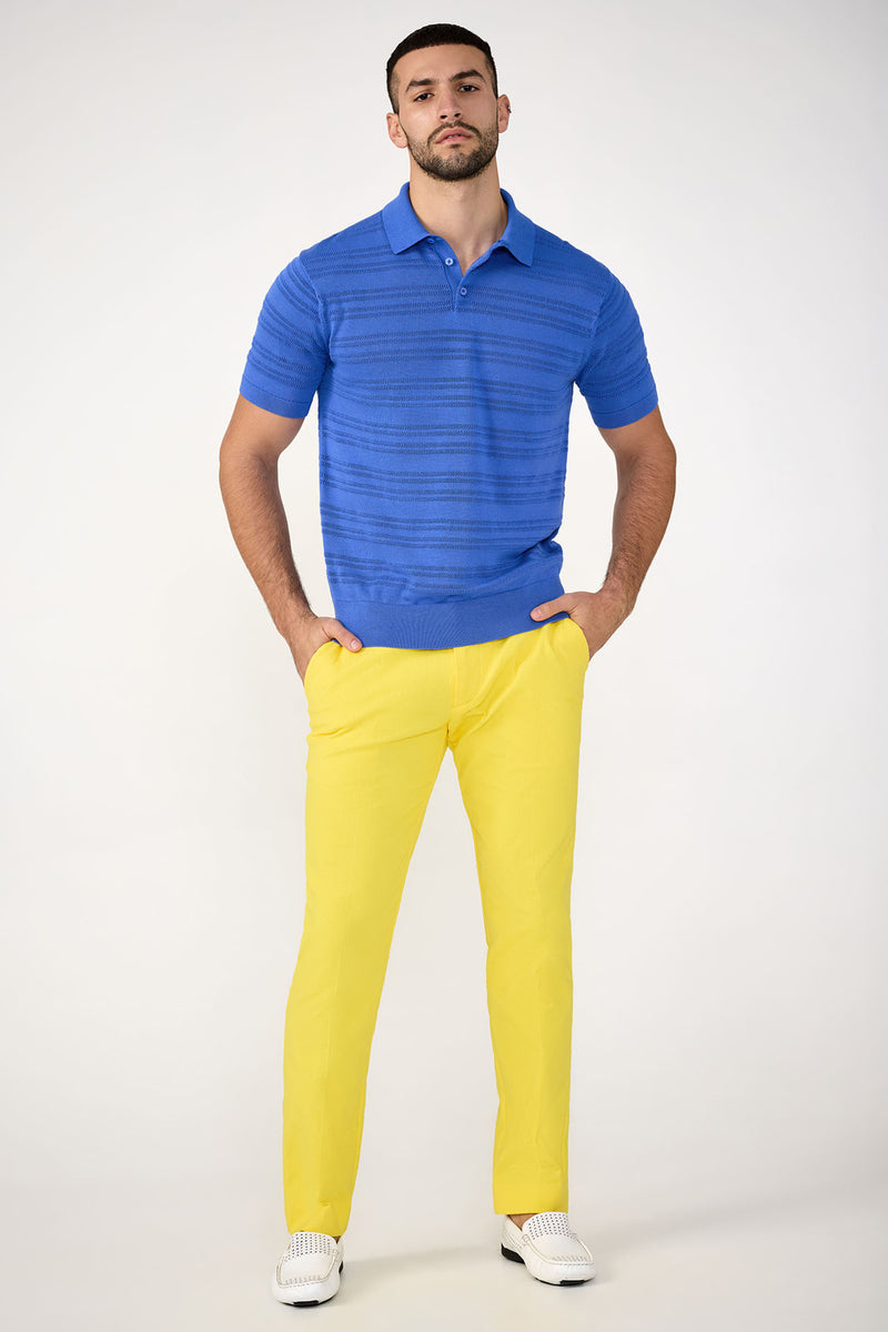 A man stands against a plain background wearing a bright blue polo, white shoes, and the Clyde Slim Trouser - Stretch Herringbone in yellow. His hands are in his pockets as he faces forward with a neutral expression.