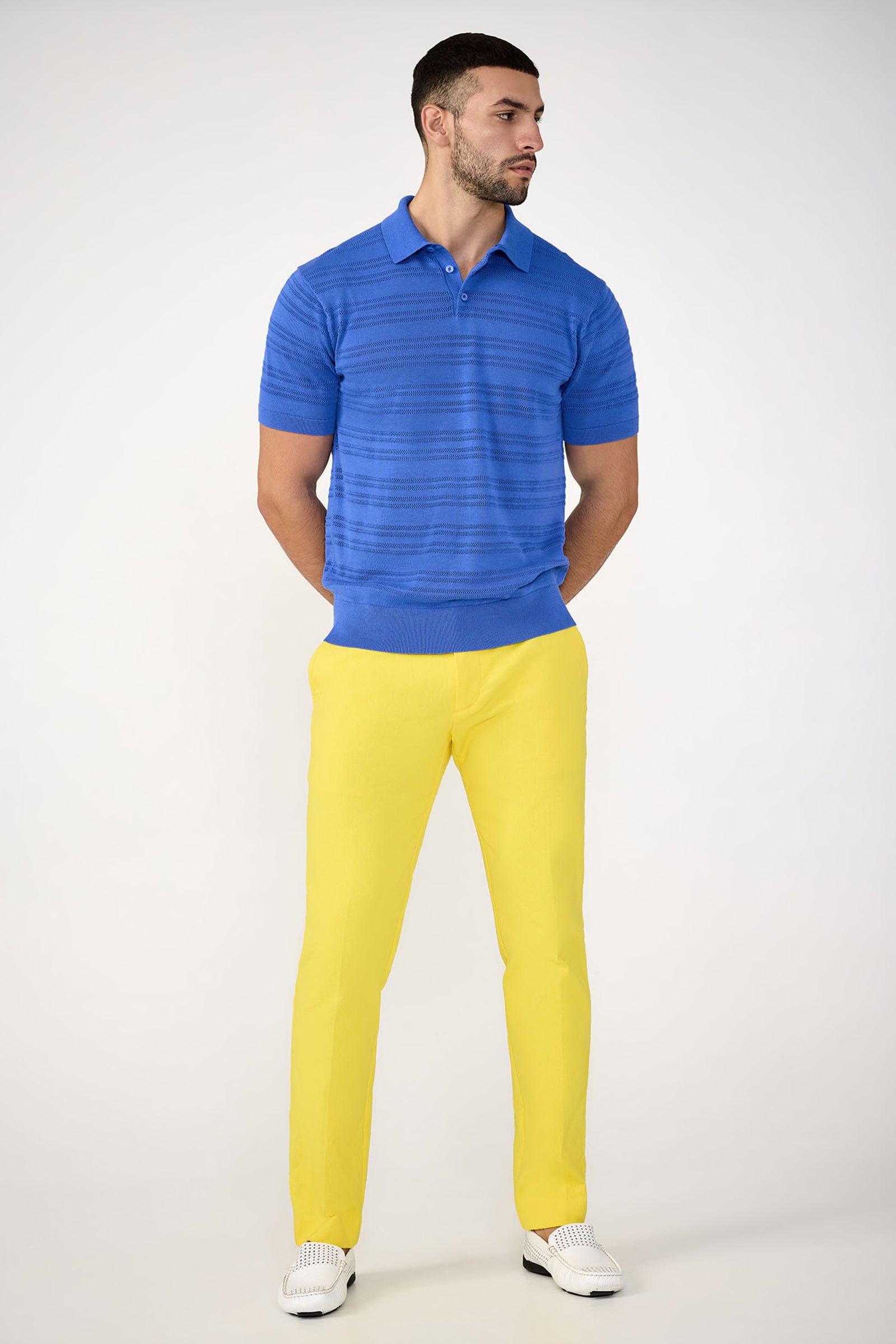A man with short dark hair and a beard stands against a plain background wearing the Edison Short-Sleeve Striped Polo, yellow pants, and white loafers, with his hands behind his back.