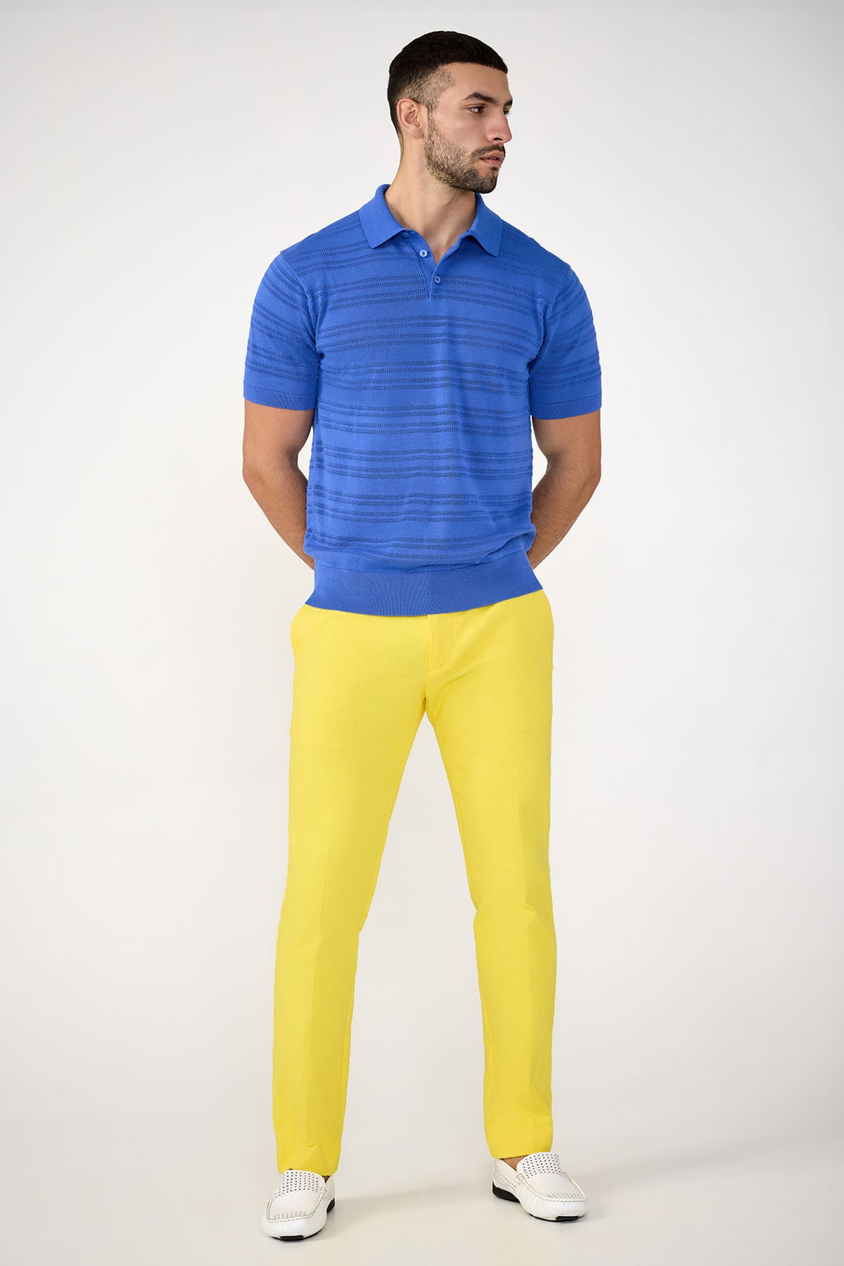 A man with short dark hair and a beard stands against a plain background wearing the Edison Short-Sleeve Striped Polo, yellow pants, and white loafers, with his hands behind his back.