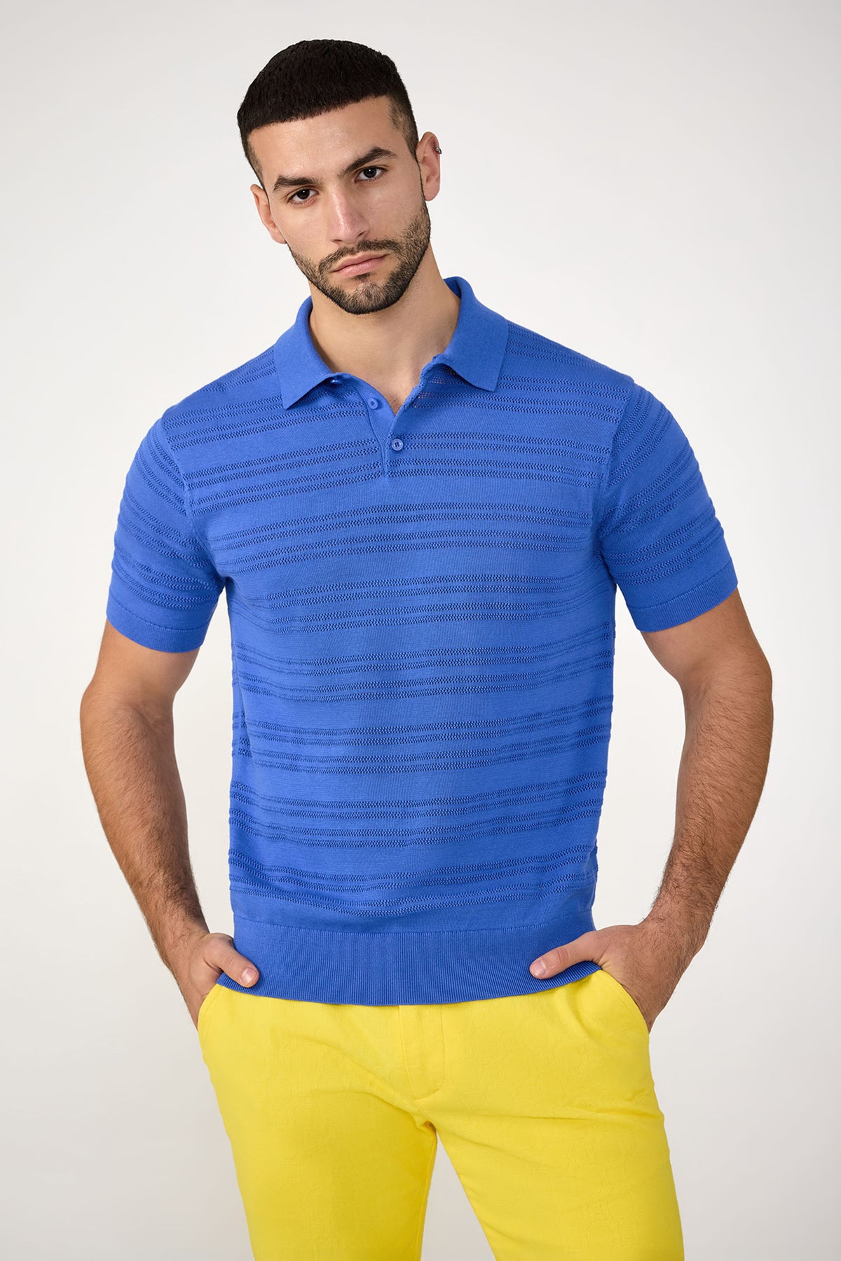 A man stands with his hands in his pockets against a plain, light background, wearing the Edison Short-Sleeve Striped Polo in Beach Ball Blue and yellow pants.