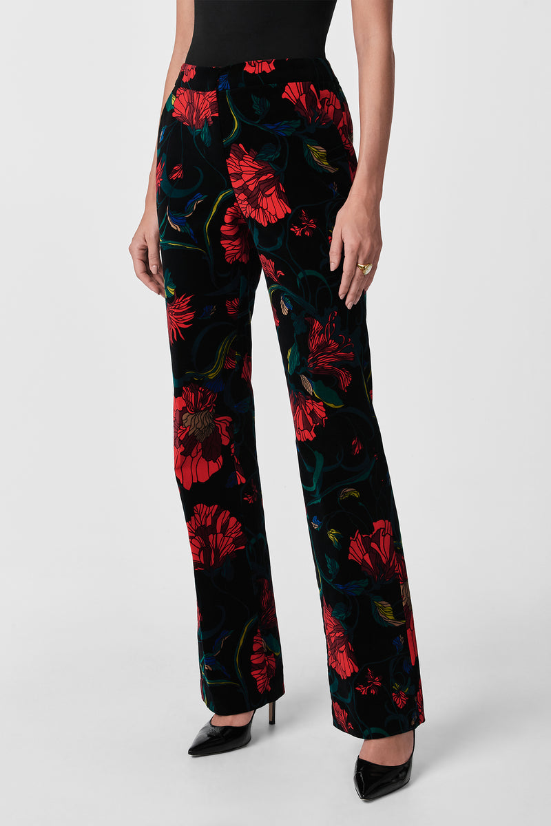 Wearing the Westport Printed Velvet Pants in a bold red floral pattern, paired with a black sleeveless top and black pointed-toe heels, this person stands against a plain light background.