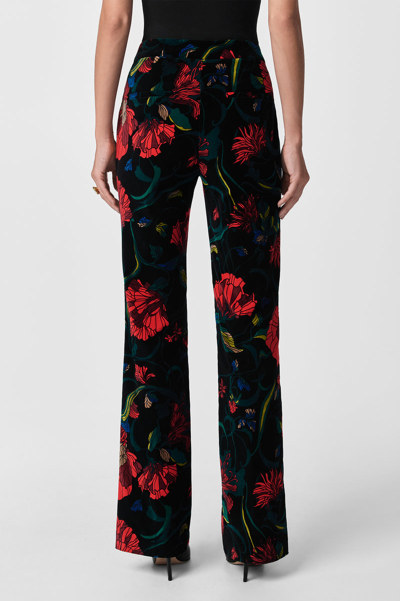 A person is seen from the back wearing Westport Printed Velvet Pants with a red and green floral design, styled with a black sleeveless top and black high-heeled shoes.
