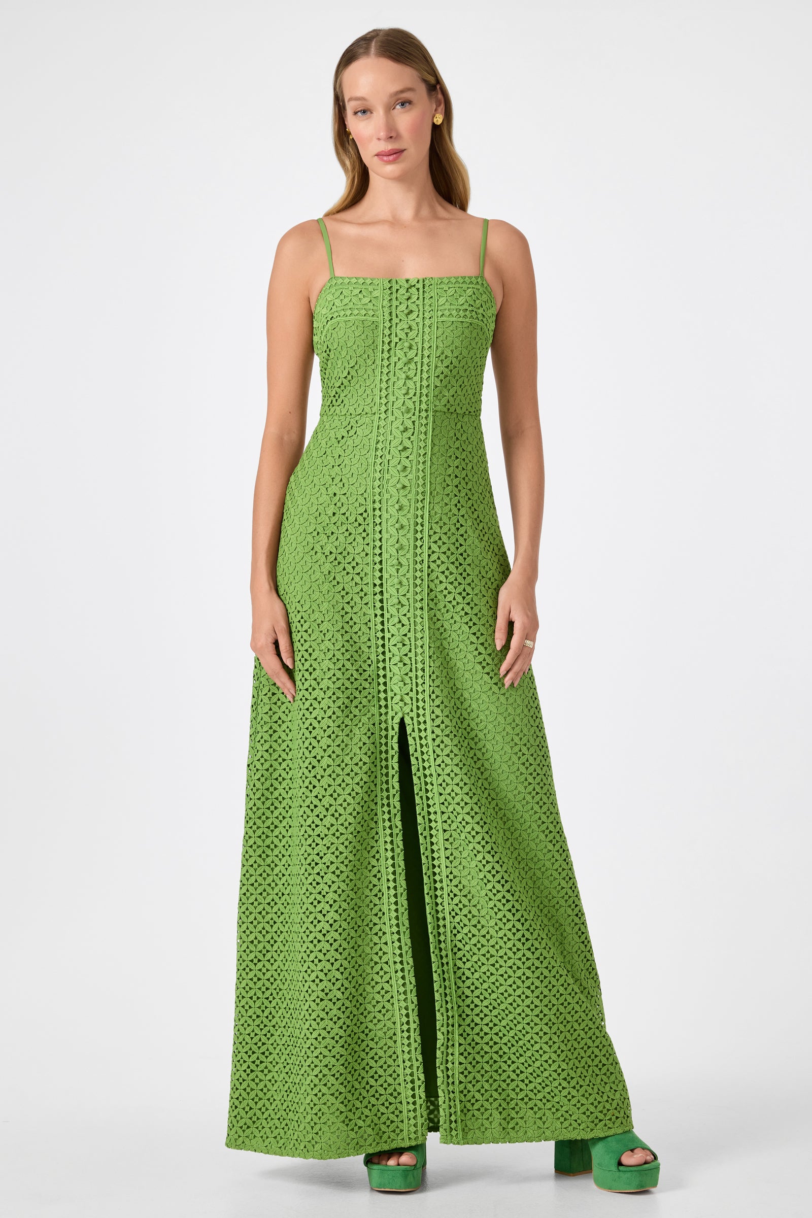 A woman wearing the Akumi Sleeveless Lace Maxi Dress in Sogen Green with thin straps and a front slit, paired with matching green platform shoes, stands against a plain white background.