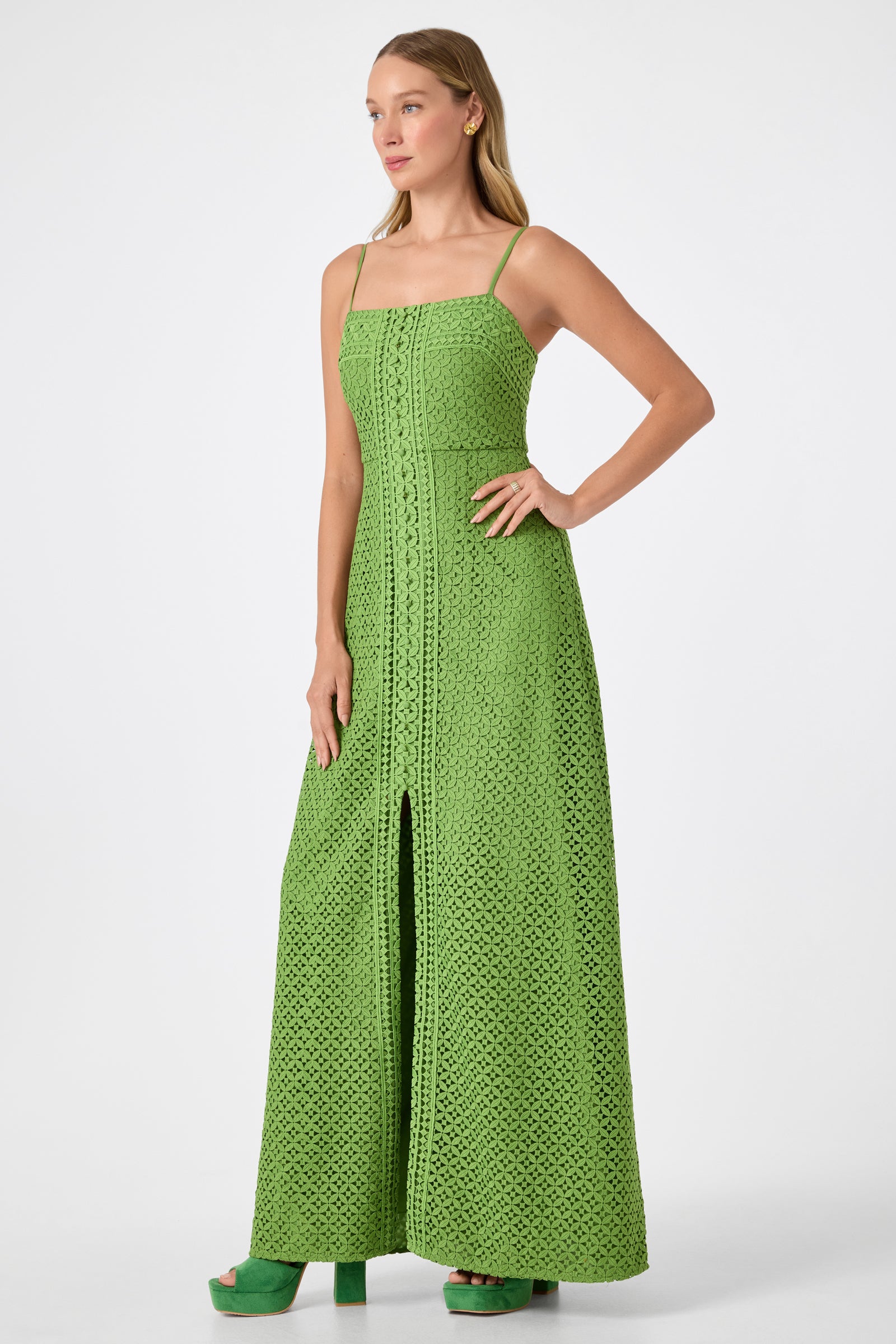 A woman poses against a plain white background wearing the Akumi Sleeveless Lace Maxi Dress, a long green crochet piece with thin straps and a front slit, paired with matching green platform heels.
