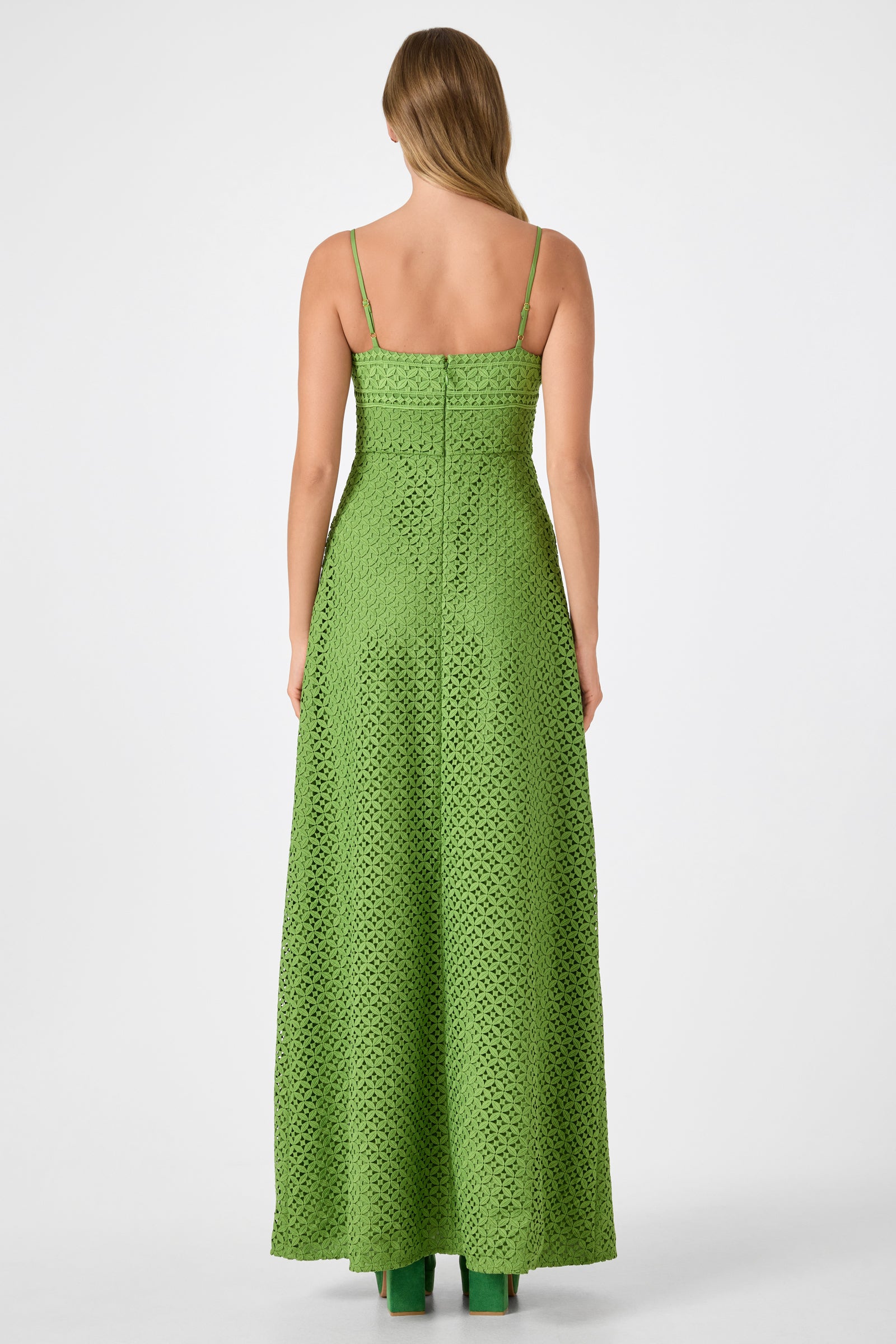 A woman with straight, light brown hair stands facing away against a white background, wearing the Akumi Sleeveless Lace Maxi Dress—a long green crochet dress with thin straps and a fitted bodice.