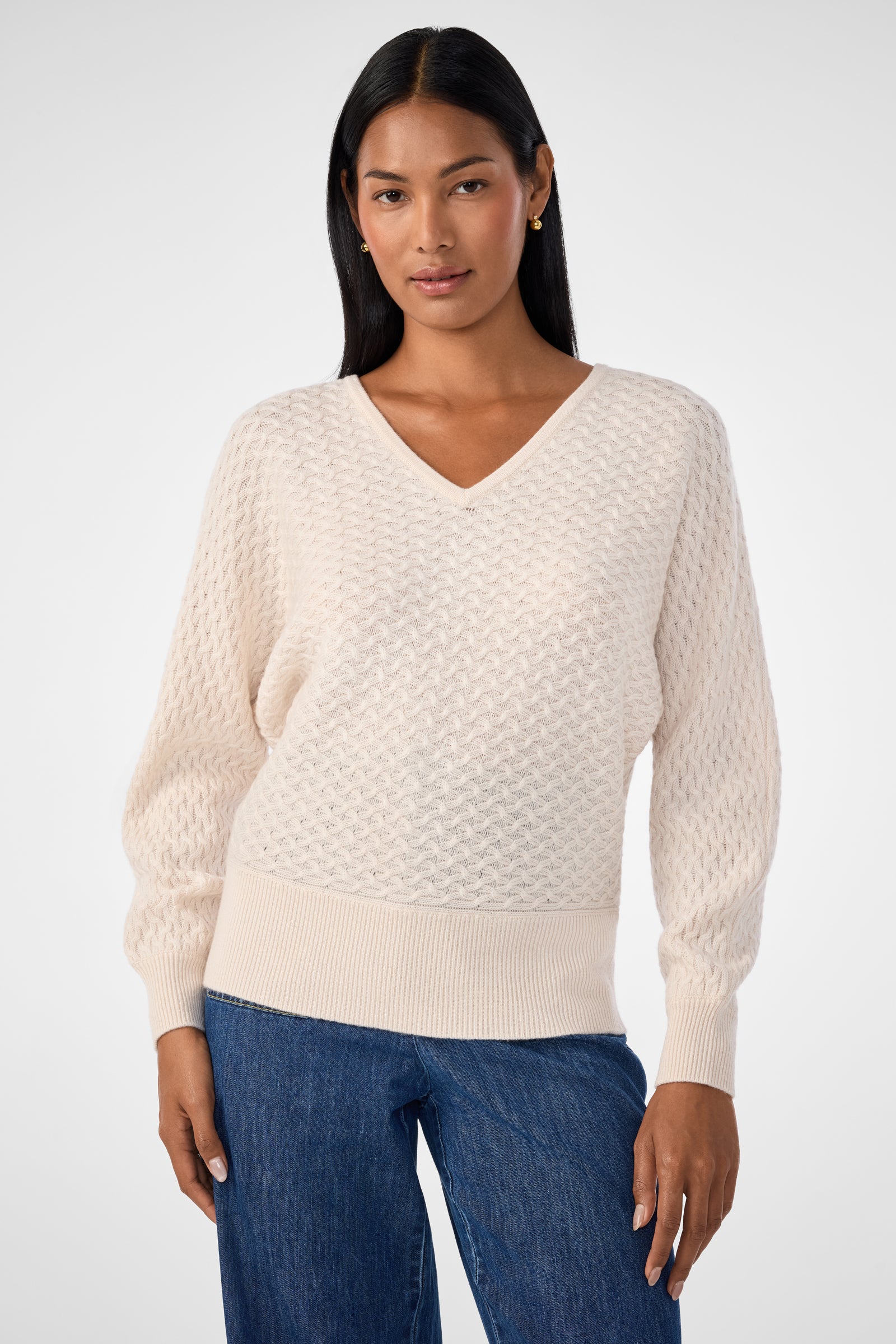 A woman with long dark hair wears the Lima V-Neck Pullover in Poplar, a textured knit sweater with long sleeves, paired with blue jeans, standing against a plain light gray background.