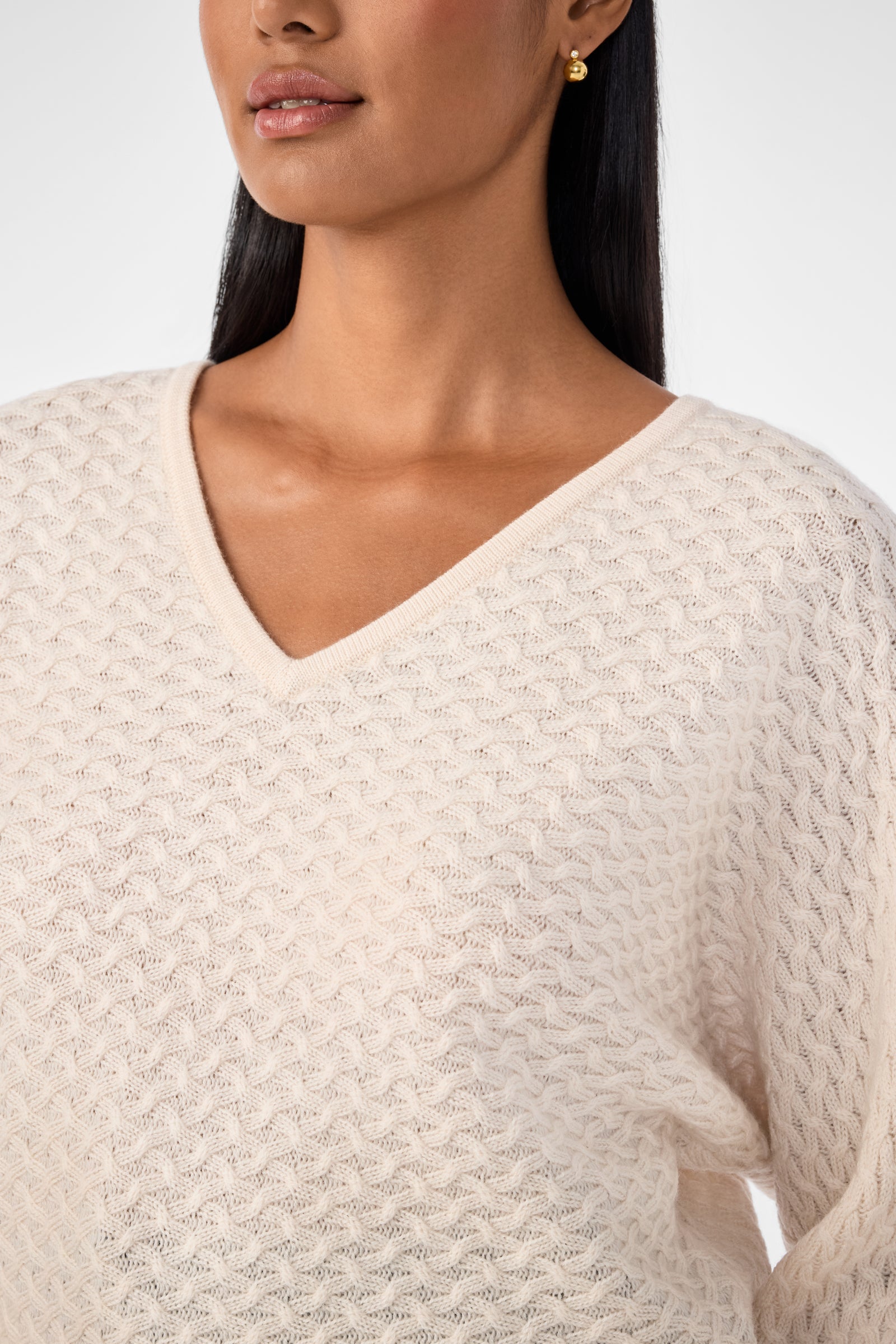 A woman wears the Lima V-Neck Pullover, a textured cream sweater, paired with gold stud earrings. The image is cropped from her lips to just below her chest.