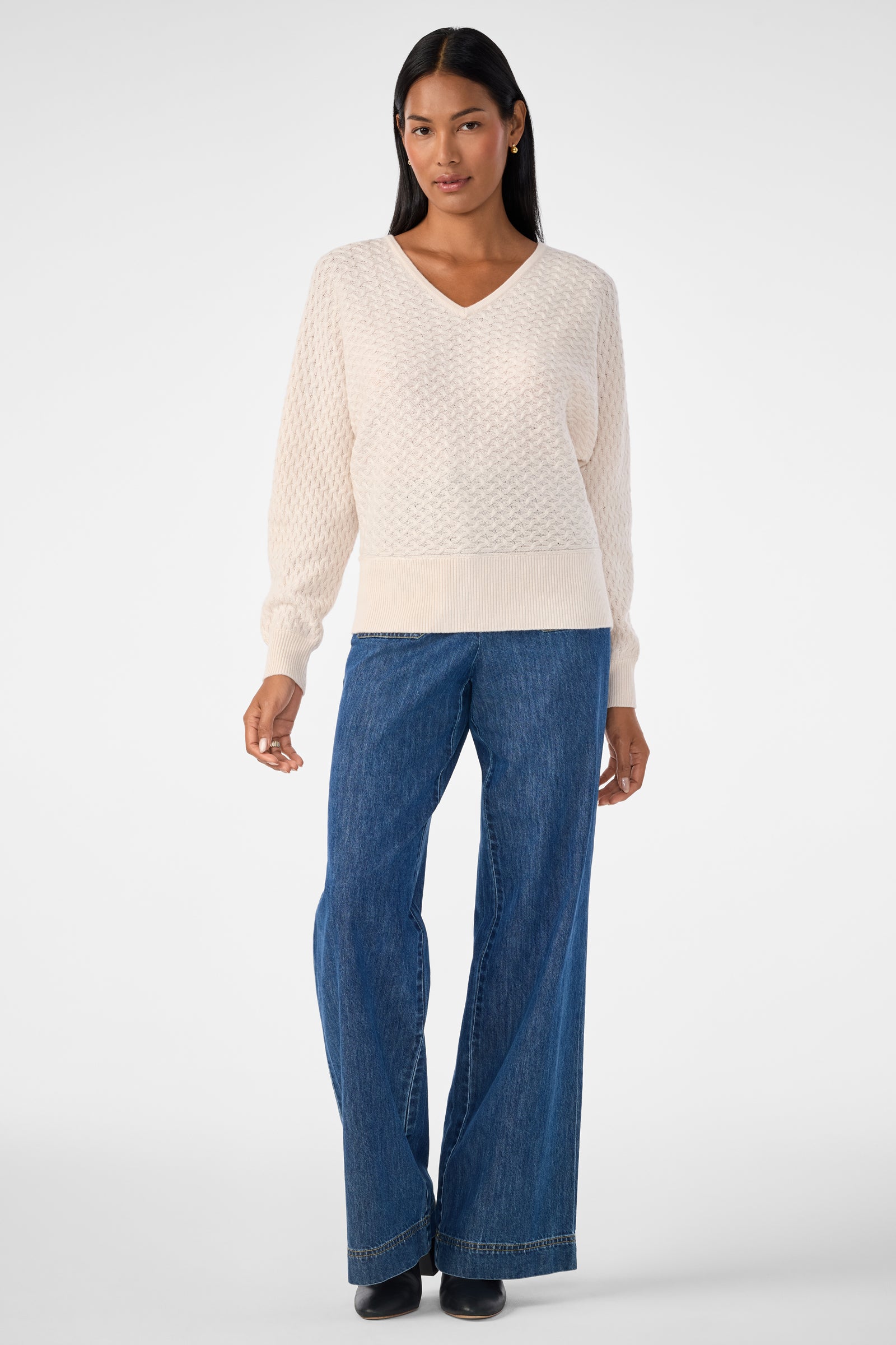 A woman with long dark hair poses against a plain white background wearing the Lima V-Neck Pullover, a cream-colored textured sweater, along with blue wide-leg jeans and black shoes. She keeps her jewelry minimal for a clean look.