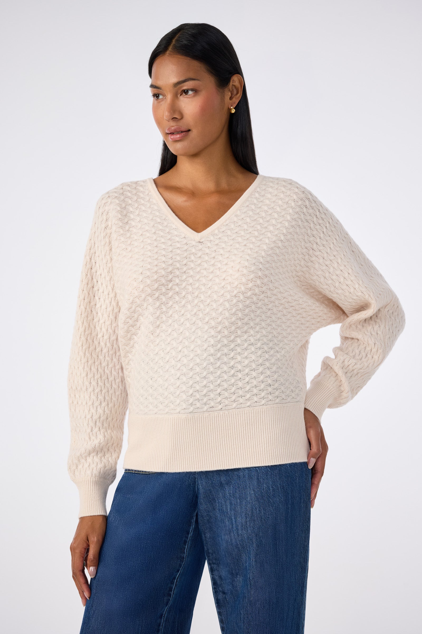 A woman wears the Lima V-Neck Pullover, a cream textured knit sweater, paired with blue jeans. She stands against a plain light background, one hand on her hip and looking slightly to the side.