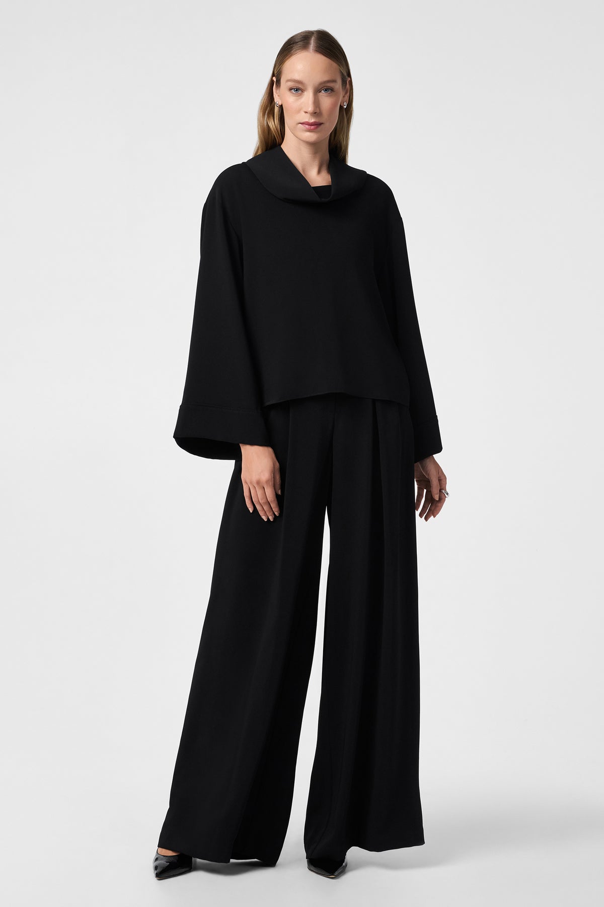 A woman stands against a plain background wearing the Staged Top, a loose black top with wide sleeves, paired with matching wide-leg black pants and black pointed-toe shoes.