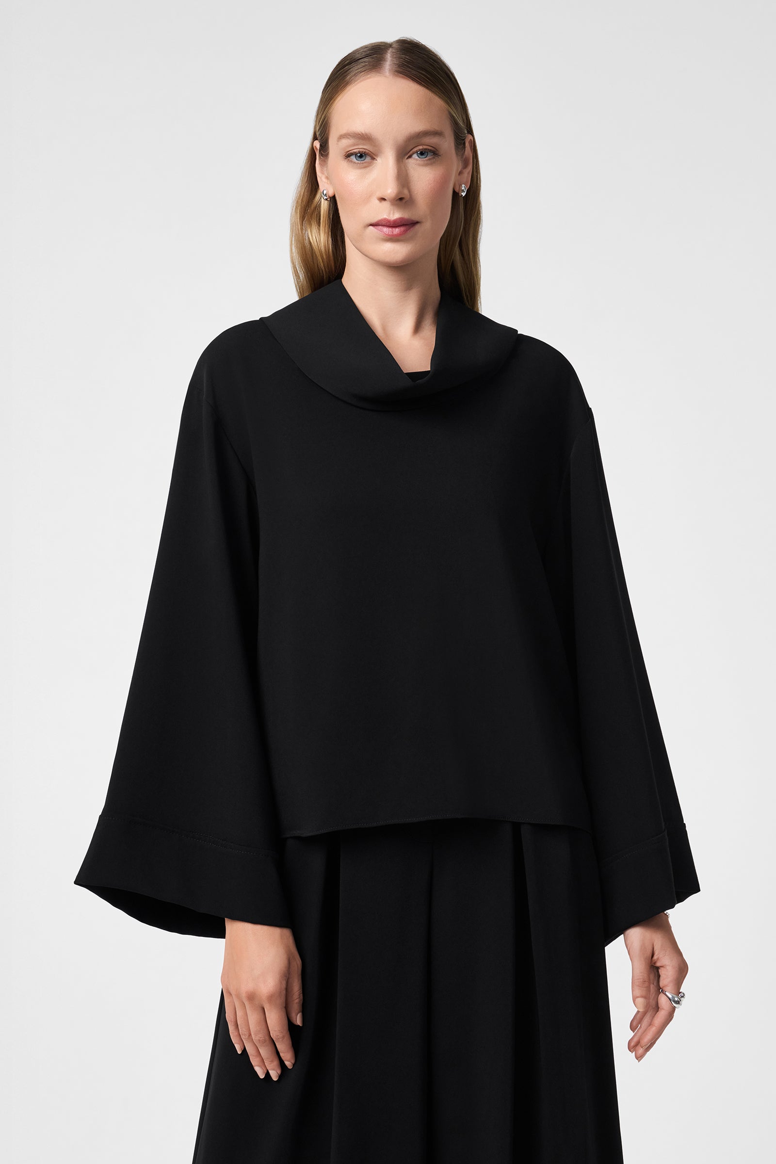 A woman with straight, light brown hair wears the loose-fitting, long-sleeved black Staged Top with a wide collar and cuffs, paired with a matching black skirt, standing against a plain light gray background.