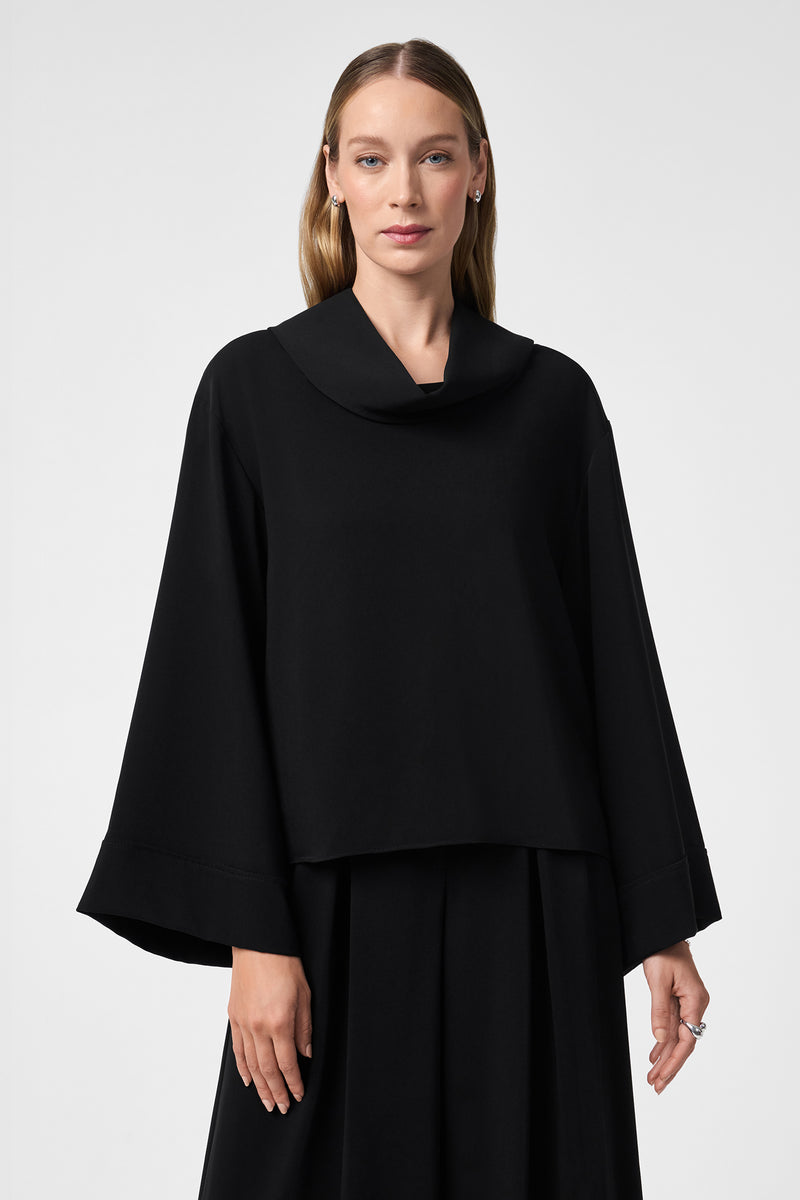 A woman with straight, light brown hair wears the loose-fitting, long-sleeved black Staged Top with a wide collar and cuffs, paired with a matching black skirt, standing against a plain light gray background.