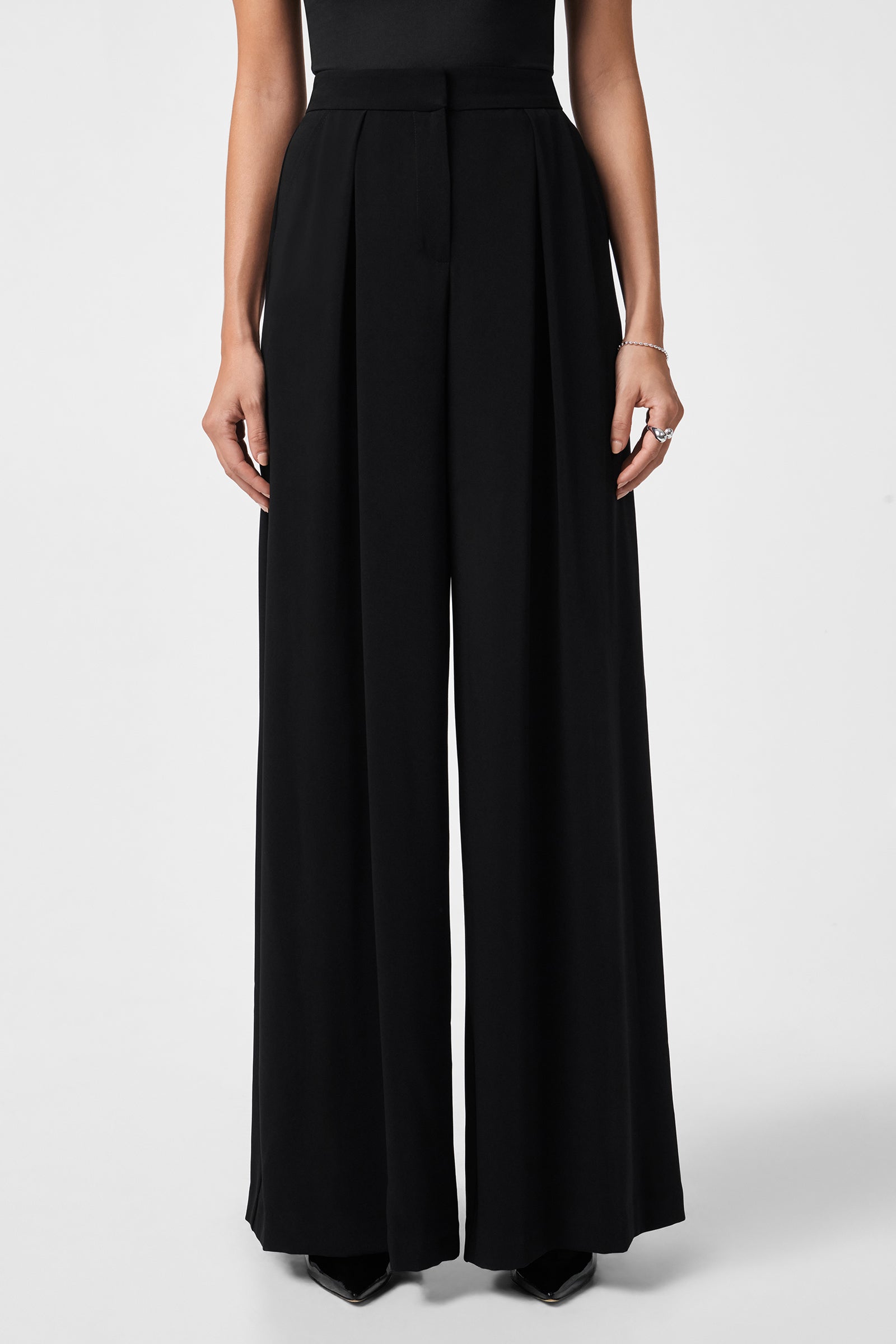 A person models the Saige Long Pant, a high-waisted, wide-leg black dress pant, paired with a sleeveless black top. Only their lower torso, legs, and hands are shown against a plain white background.